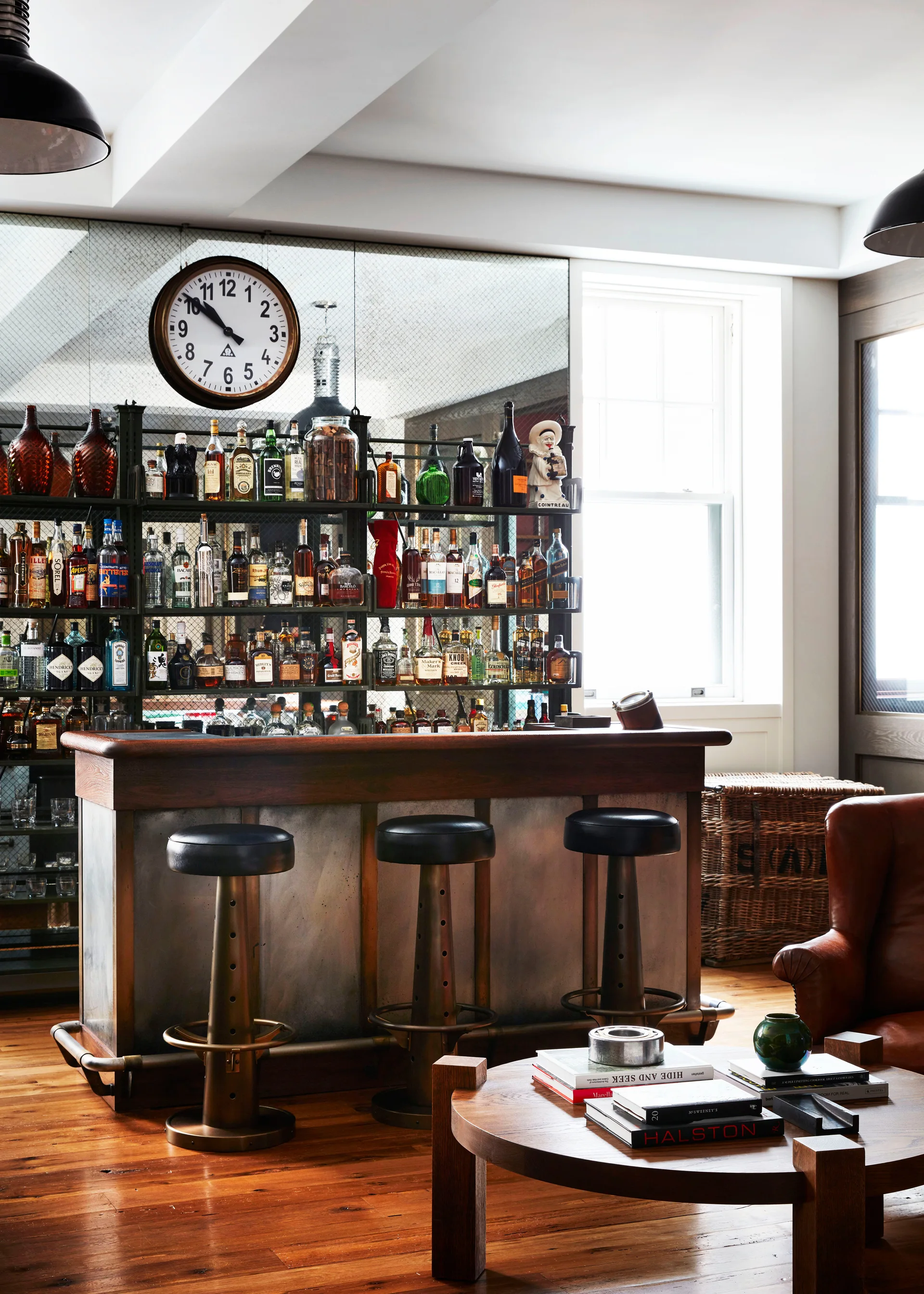 An industrial home bar features a wooden counter, three dark-topped metal stools, and a tall metal shelving unit backed by a large mirror.