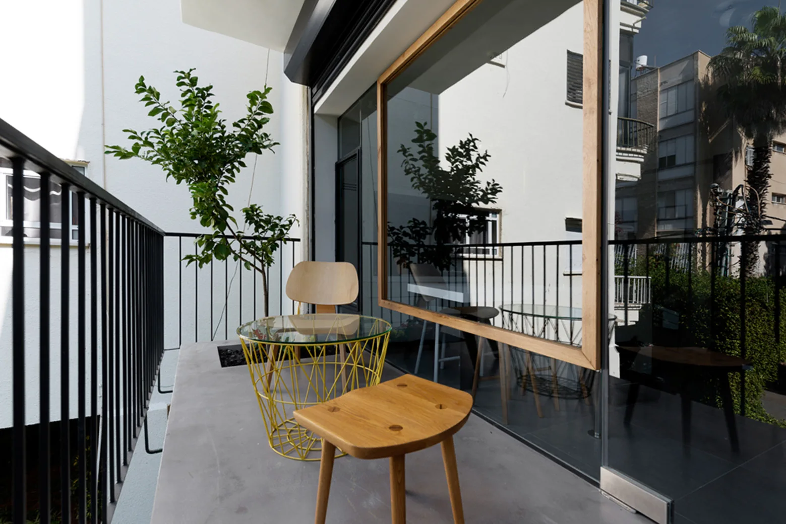 A modern apartment balcony features a yellow wire-frame table with a glass top, a wooden stool, a wooden chair, and black railings.