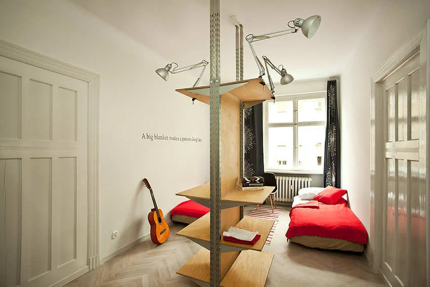 A narrow bedroom with herringbone floors, white walls, beds with red blankets, and a central wood and metal shelving unit with swing-arm lamps.