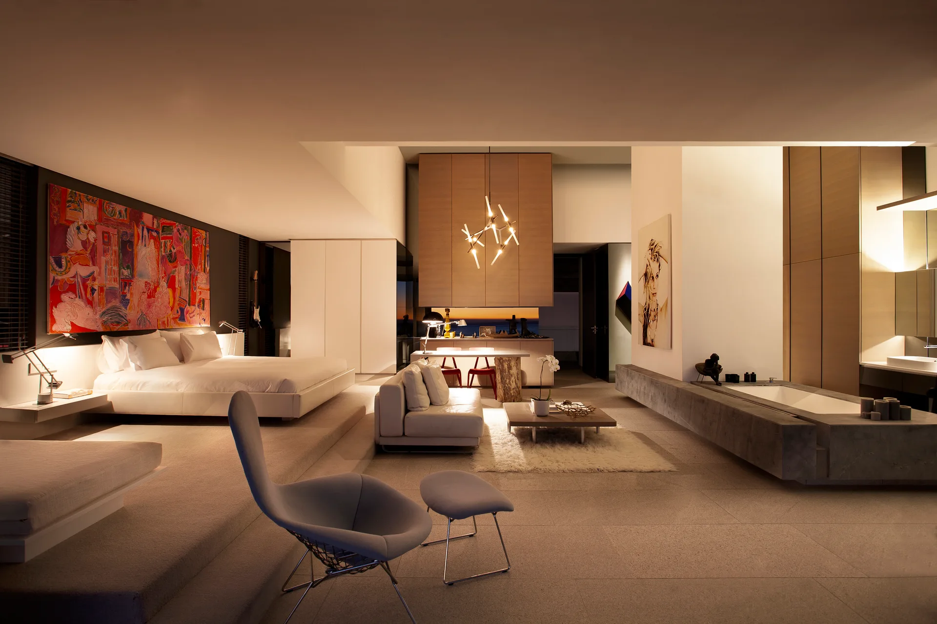 A multi-level bedroom suite featuring a raised platform bed, a sunken lounge with a white sofa, and an integrated stone bathtub under warm lighting.