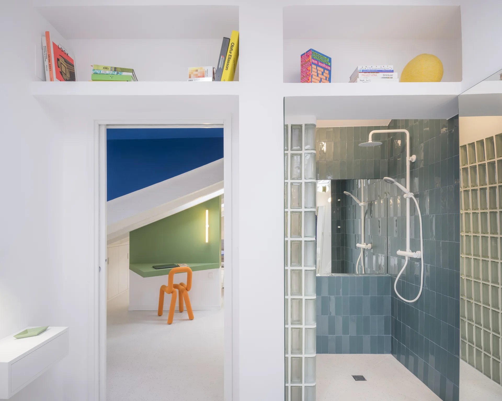 A teal tiled shower with a glass block wall sits next to a doorway looking into a white room with a green desk and orange chair.