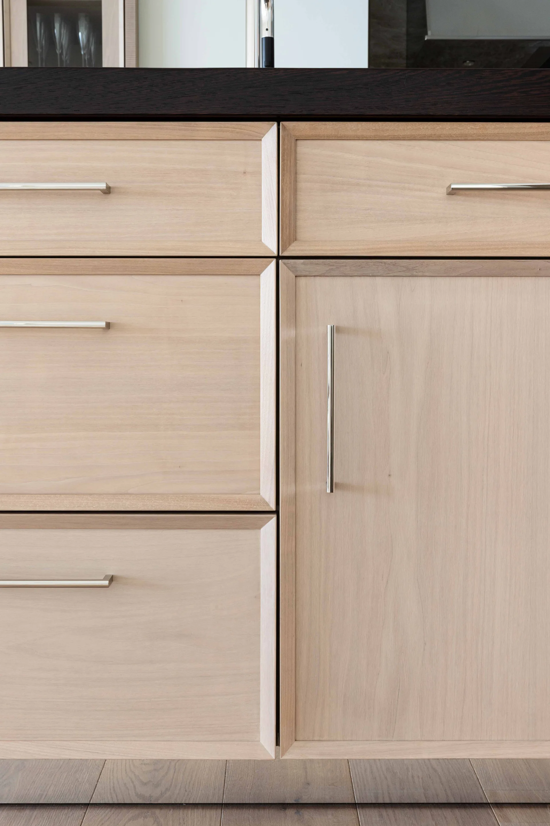 Light wood kitchen cabinets feature flat-panel drawers and doors with beveled frame borders, slim metal bar handles, and a dark countertop edge above.