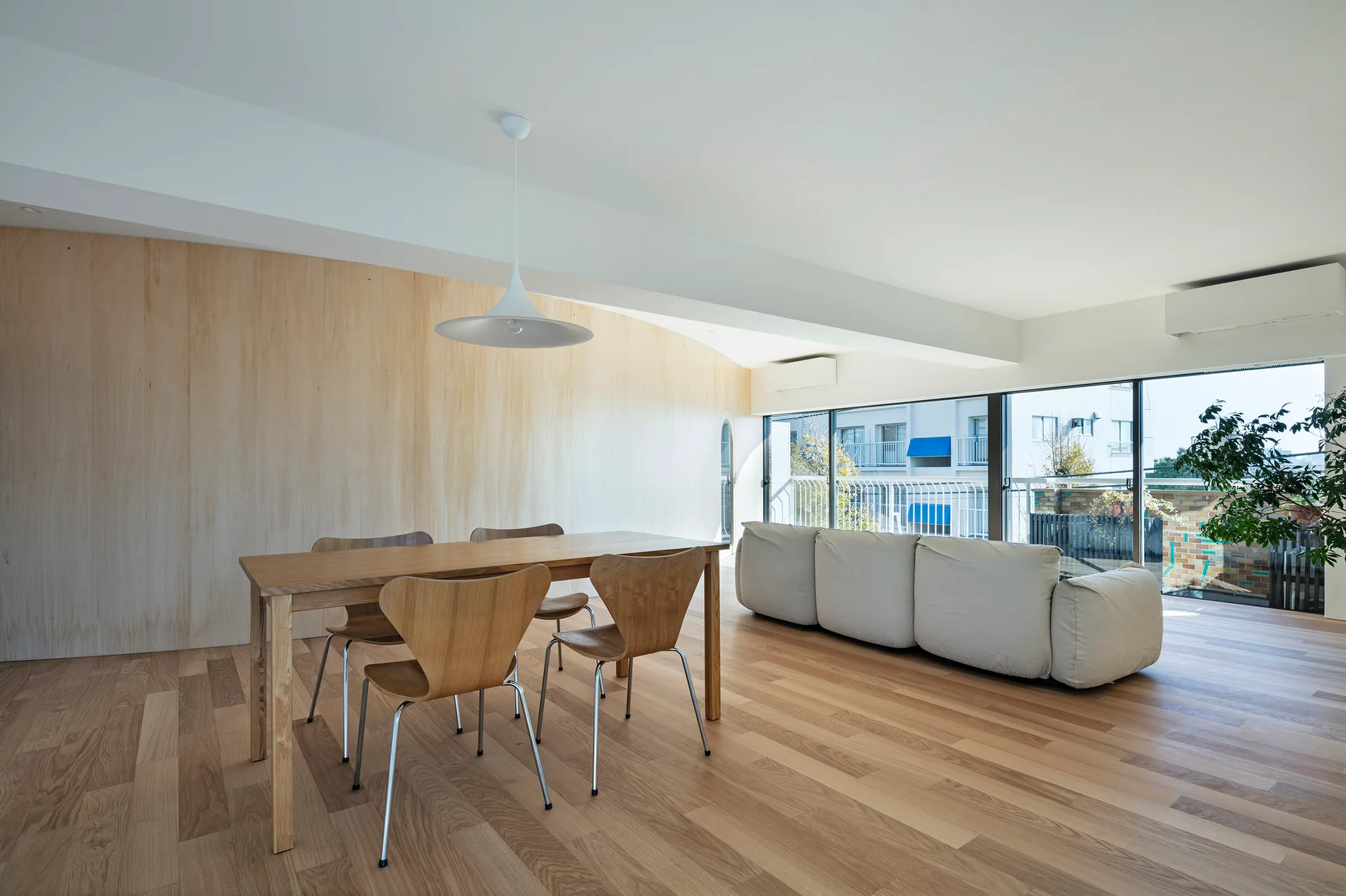 An open-plan dining and living room features a light wood paneled wall, a wooden dining table with curved chairs, and a low white sofa near large windows.