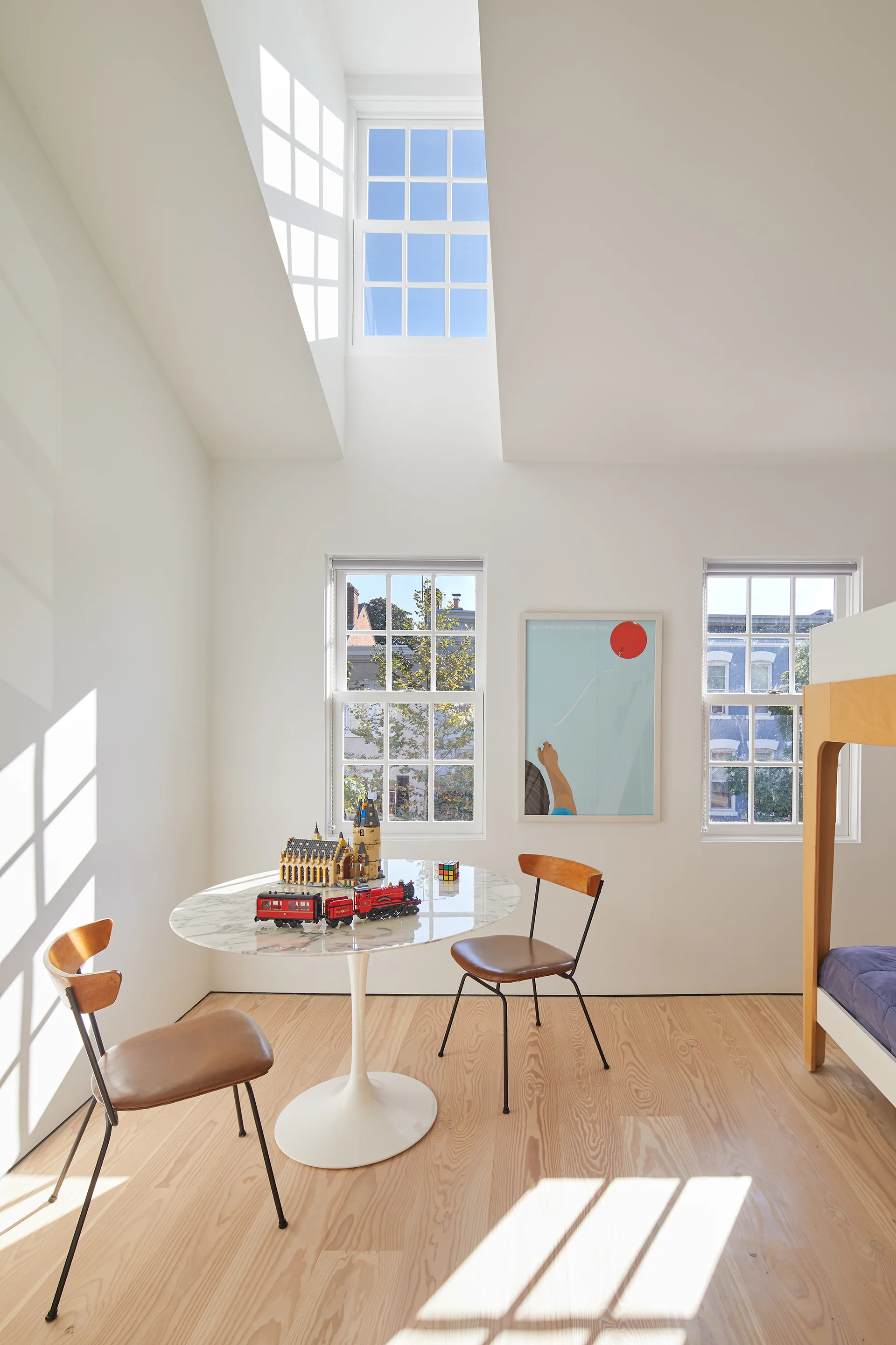 A bright kids bedroom features a white round tulip table with two chairs, two windows, and sunlight streaming down from an overhead skylight.