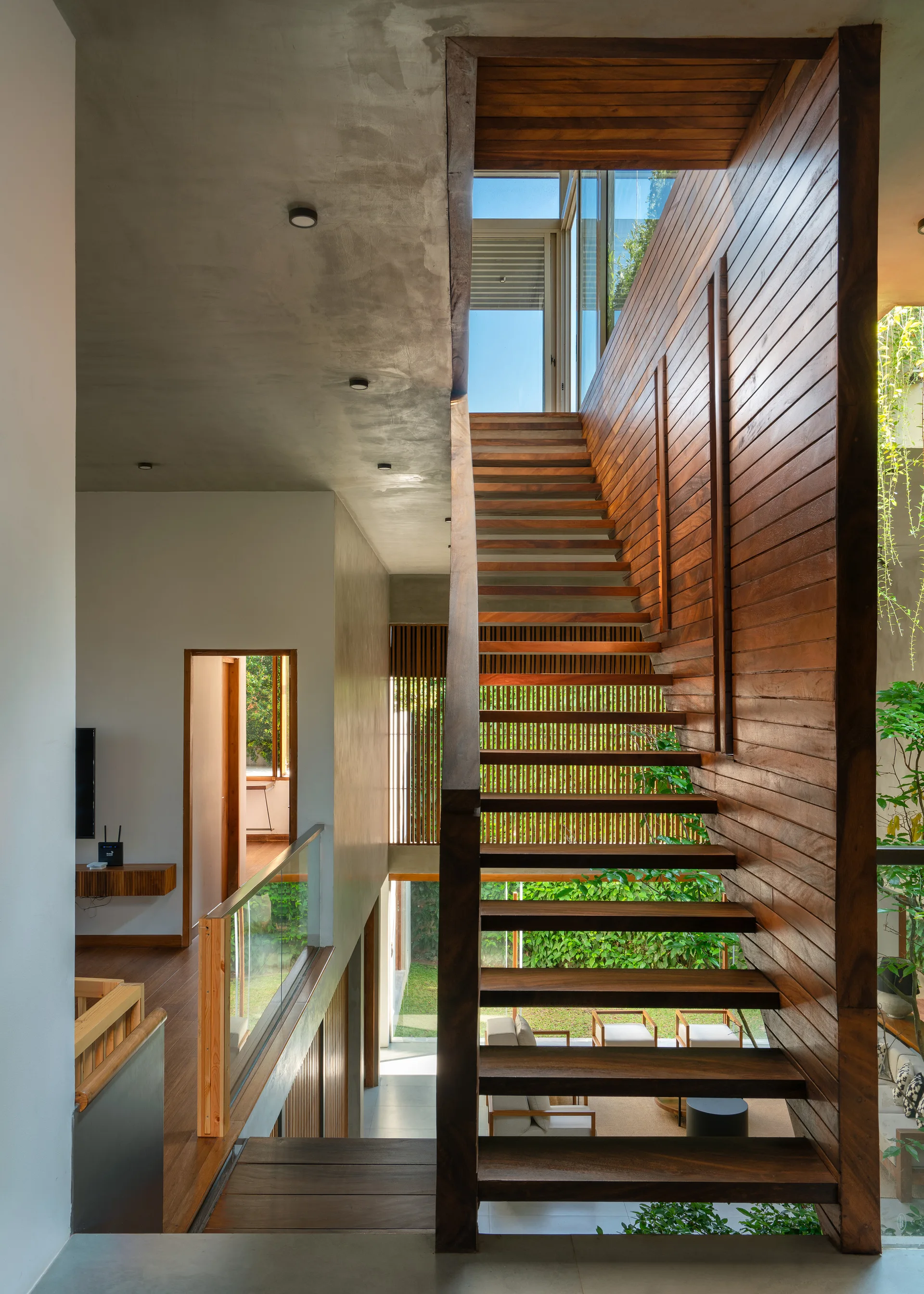A modern residential interior features a central wooden floating staircase with an exposed concrete ceiling, glass railings, and natural light from above.