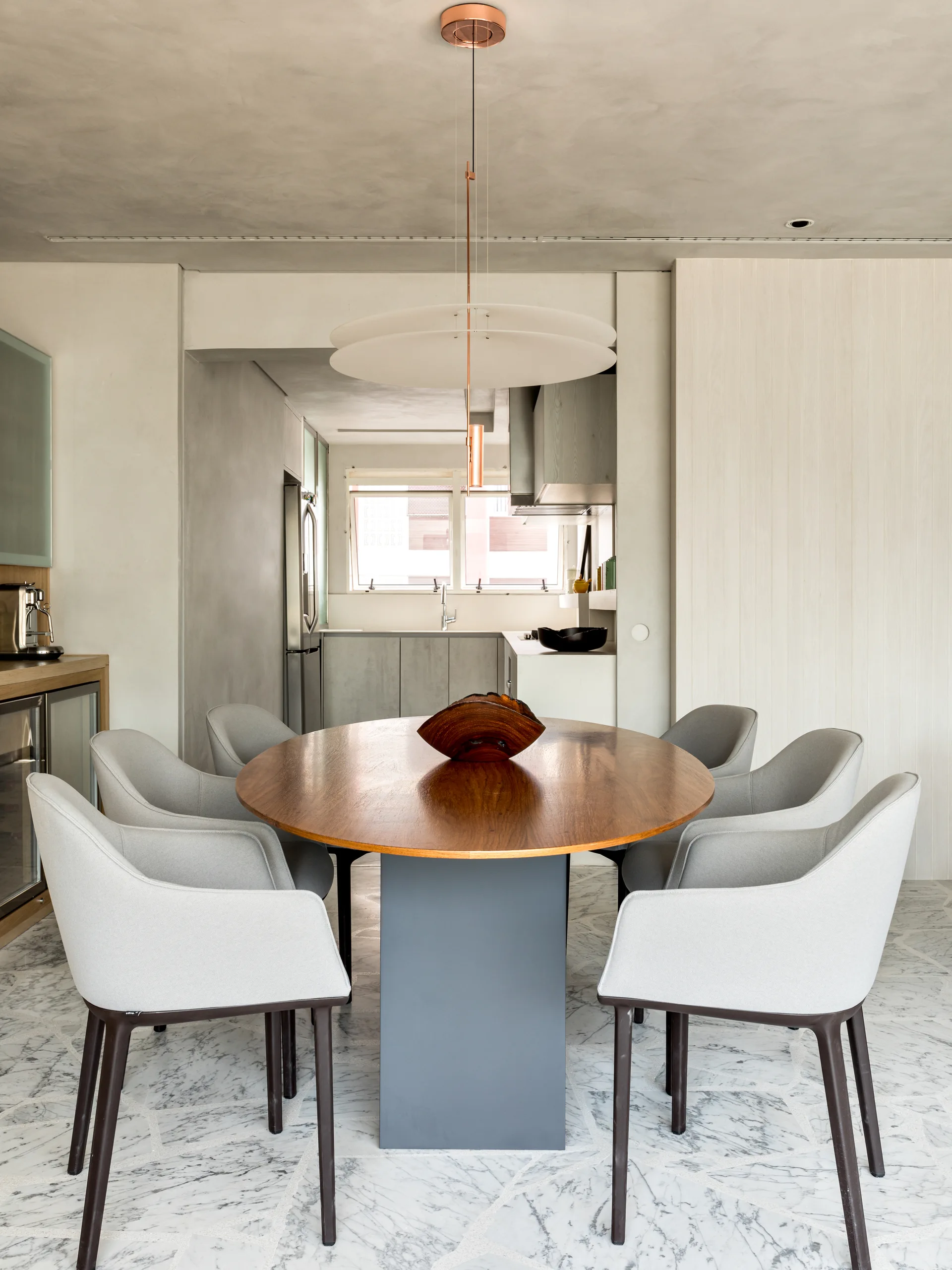 A modern dining room features a round wood table with six gray upholstered chairs on marble tile flooring beneath a large white disc pendant light.
