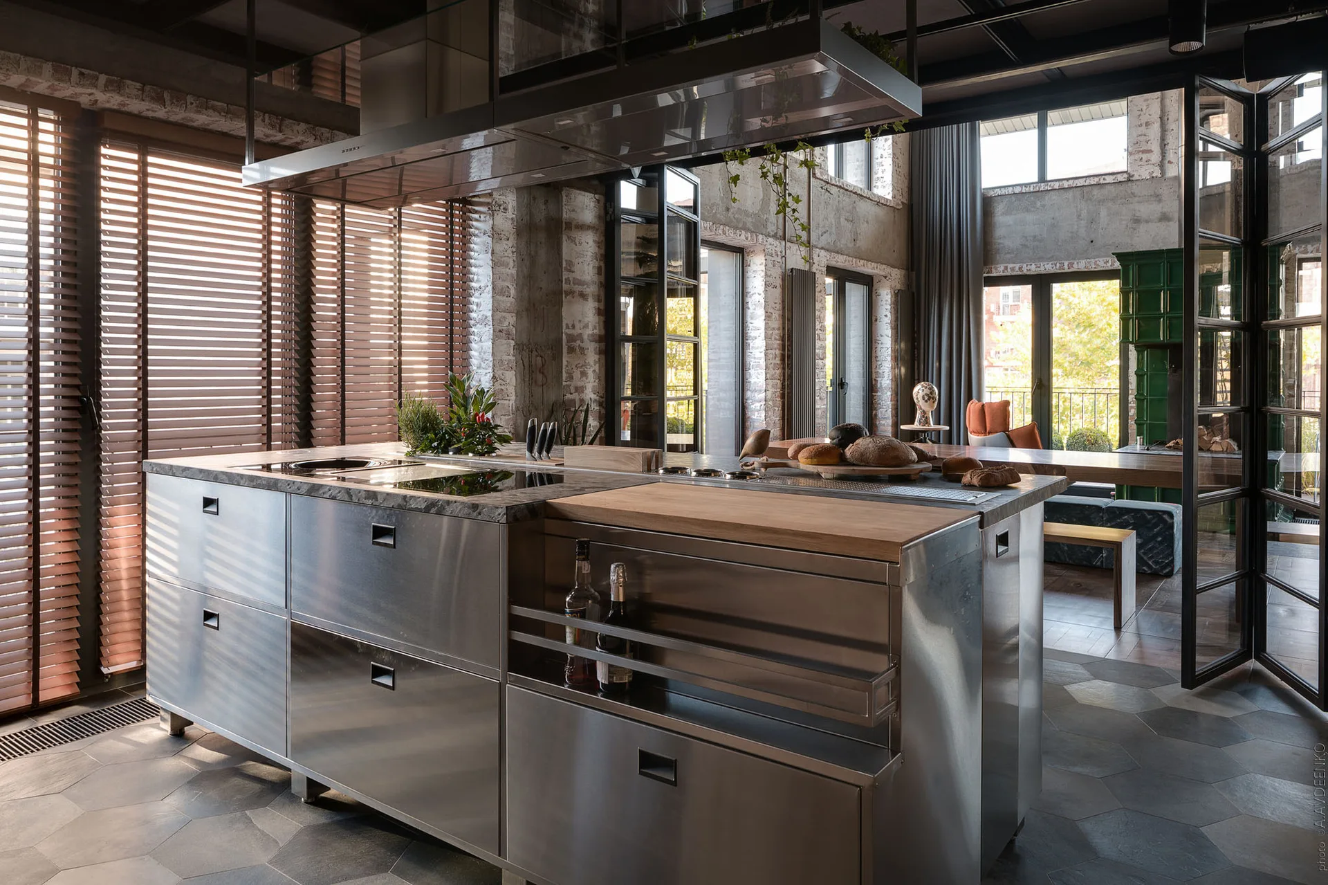 A spacious industrial loft kitchen featuring a brushed stainless steel island, grey hexagonal floor tiles, and exposed brick walls.