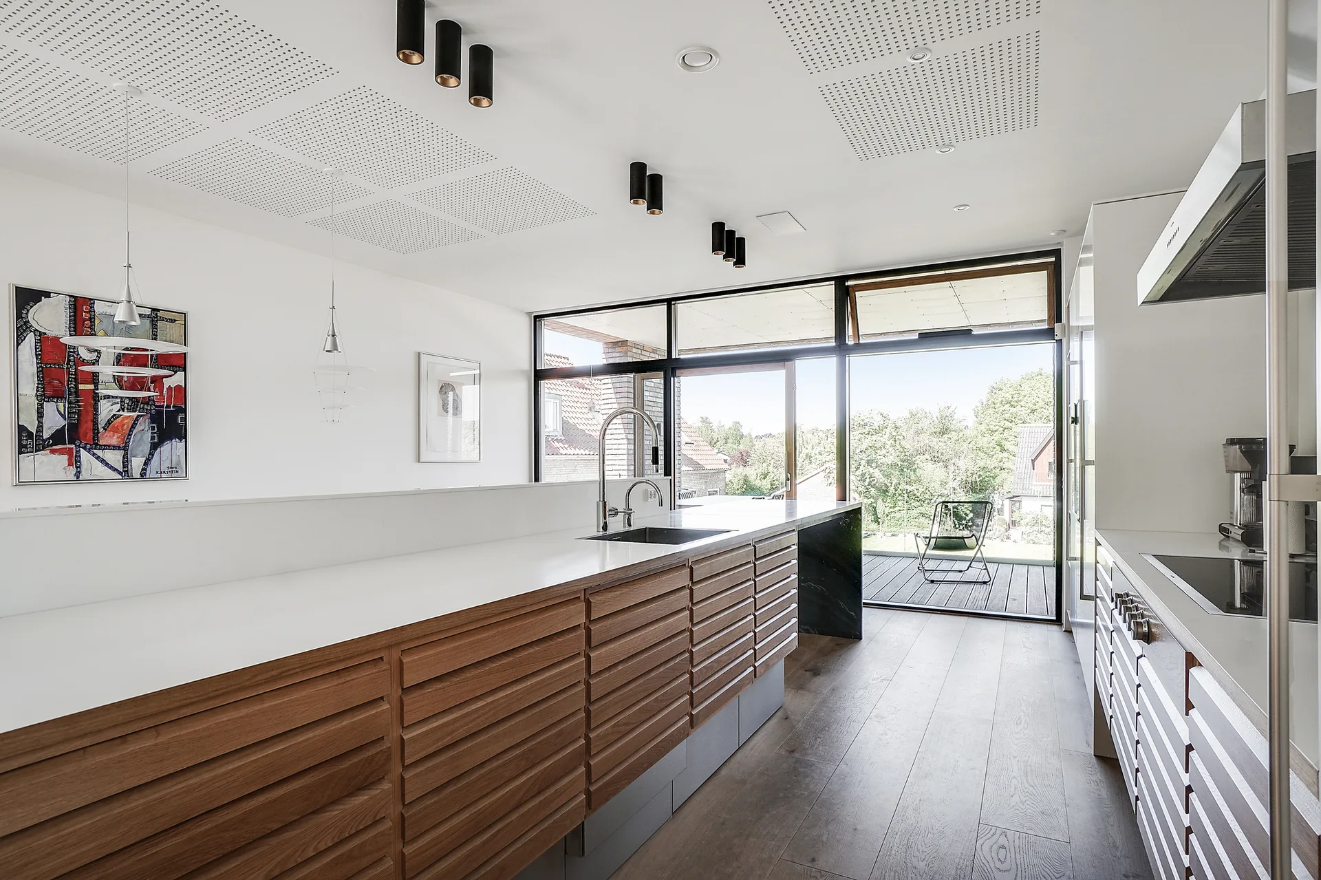 A modern kitchen featuring horizontal slatted wood cabinets, white countertops, black cylindrical ceiling lights, and a glass wall opening to a deck.