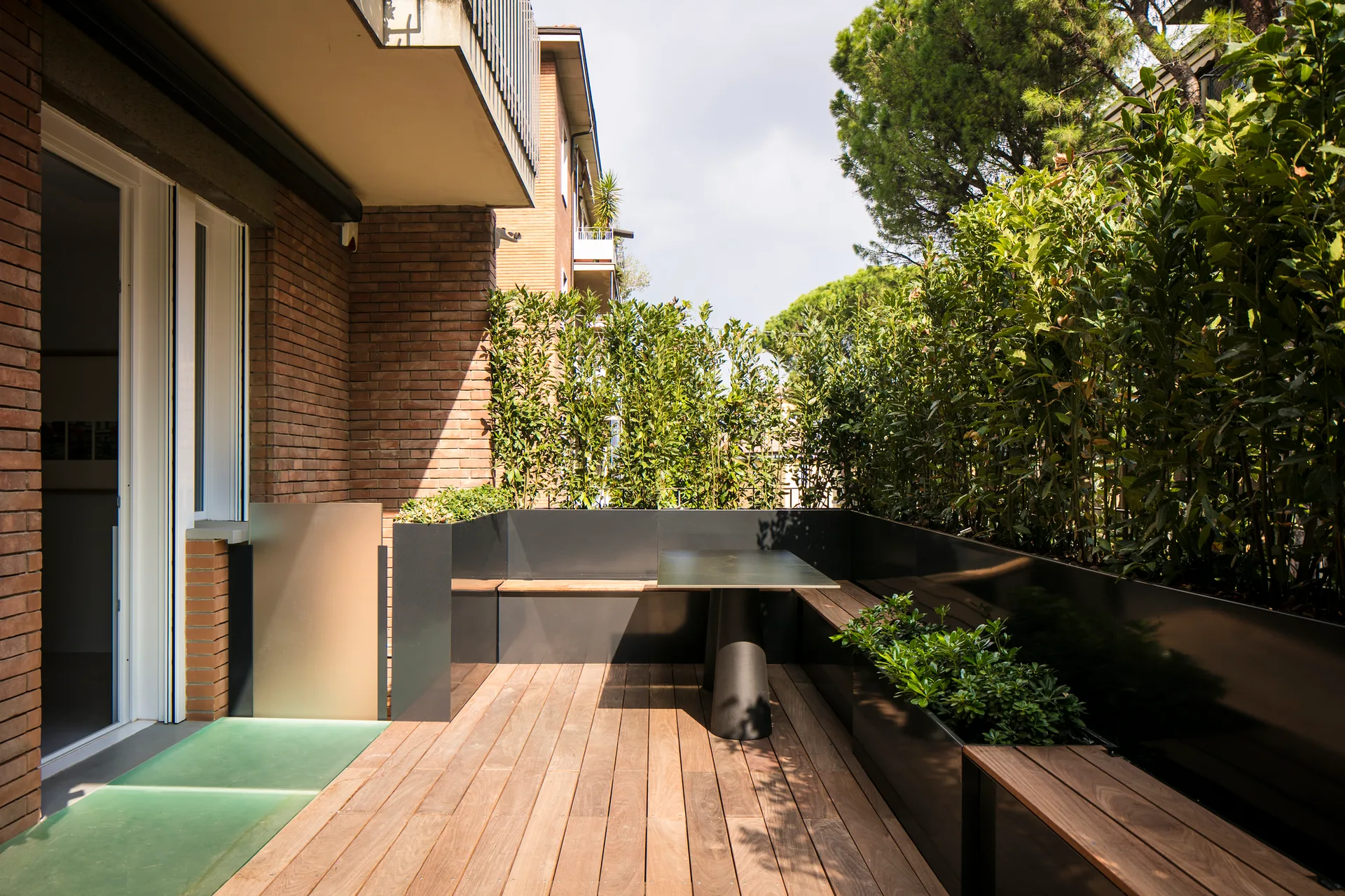 A modern outdoor terrace with wood decking, a black metal L-shaped built-in bench, a matching table, and a tall green privacy hedge.