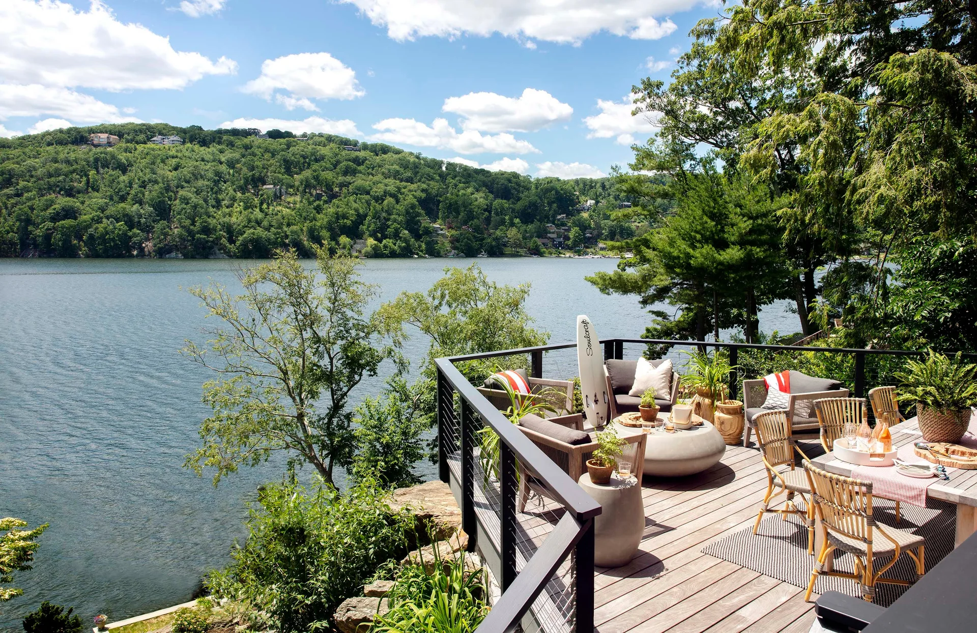 A wooden deck overlooking a lake features woven lounge chairs around a round concrete coffee table and a nearby dining area.