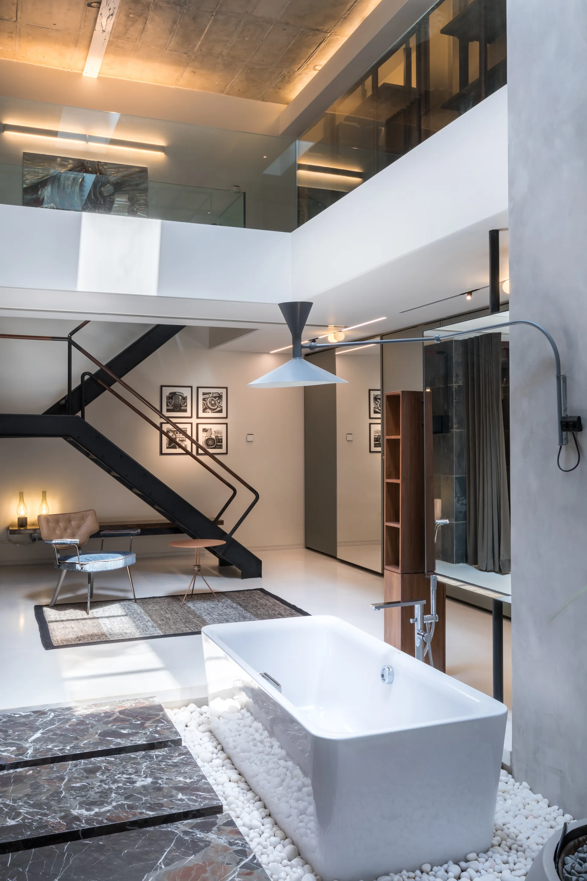A white freestanding bathtub sits on a bed of white river pebbles next to a black metal staircase in a double-height loft bathroom.