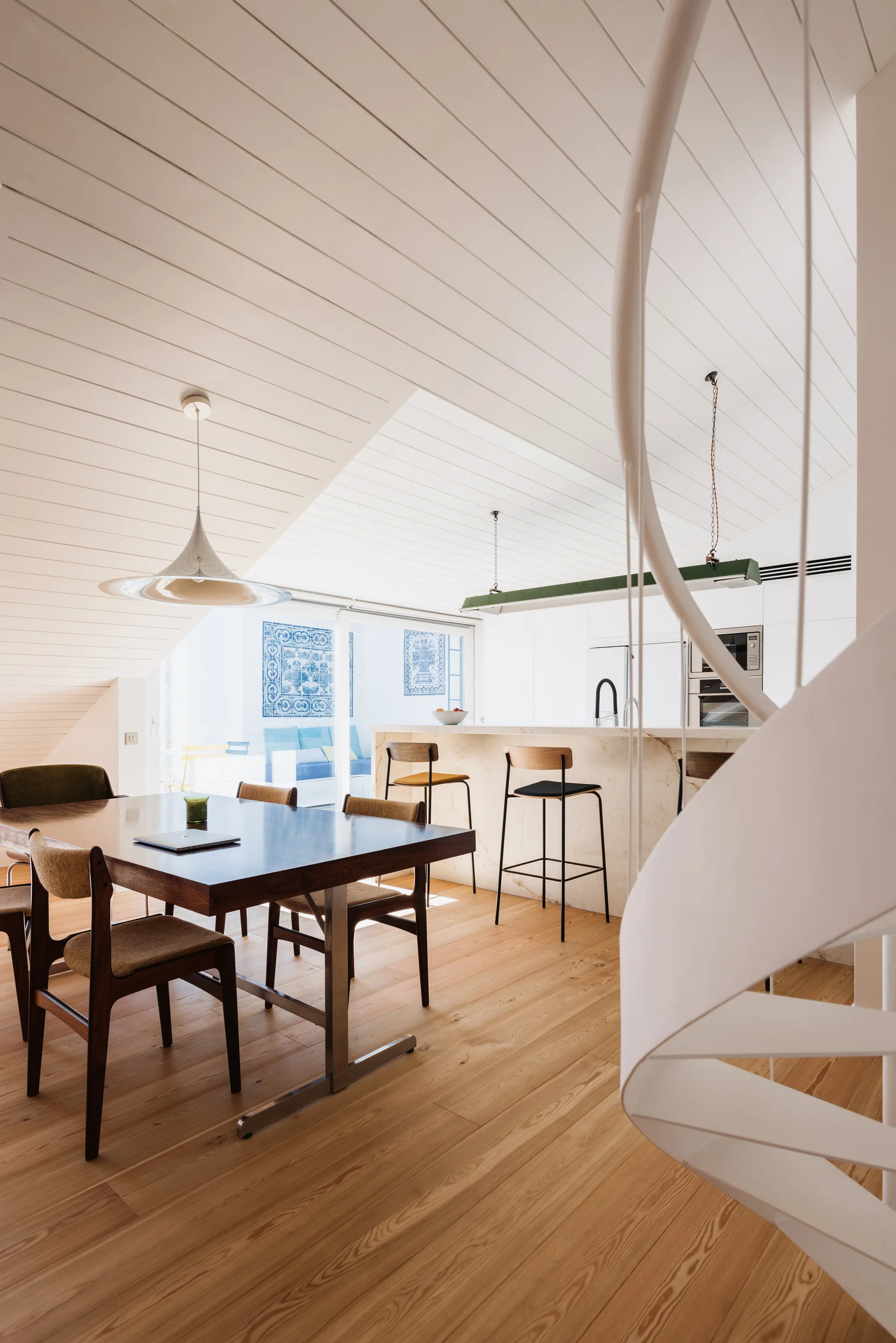 A bright dining room features a wooden table and chairs beneath a sloped white-planked ceiling, next to a white spiral staircase and kitchen island.