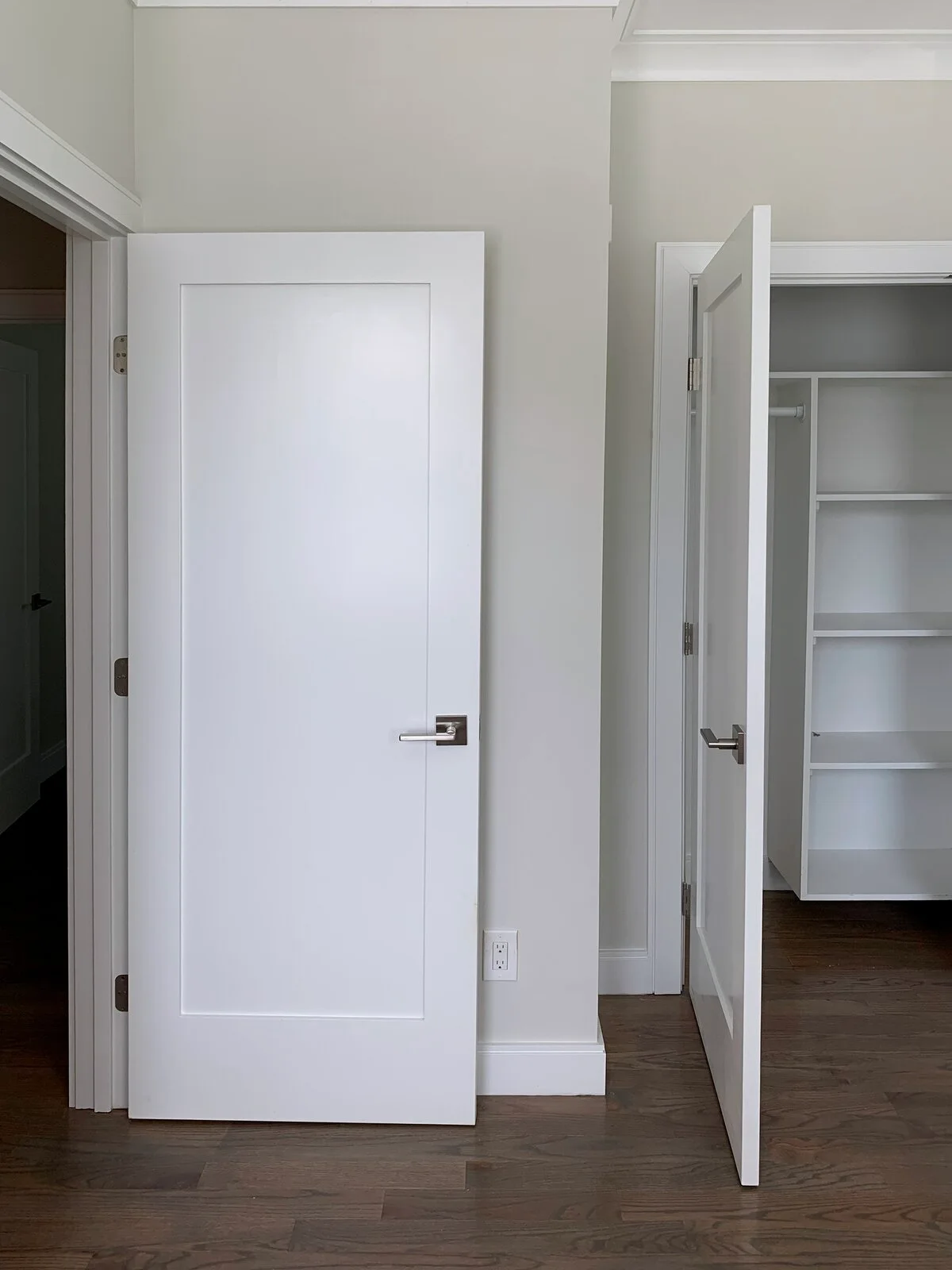 Two white shaker doors on a light greige wall, with one door open to reveal white closet shelves over dark wood flooring.