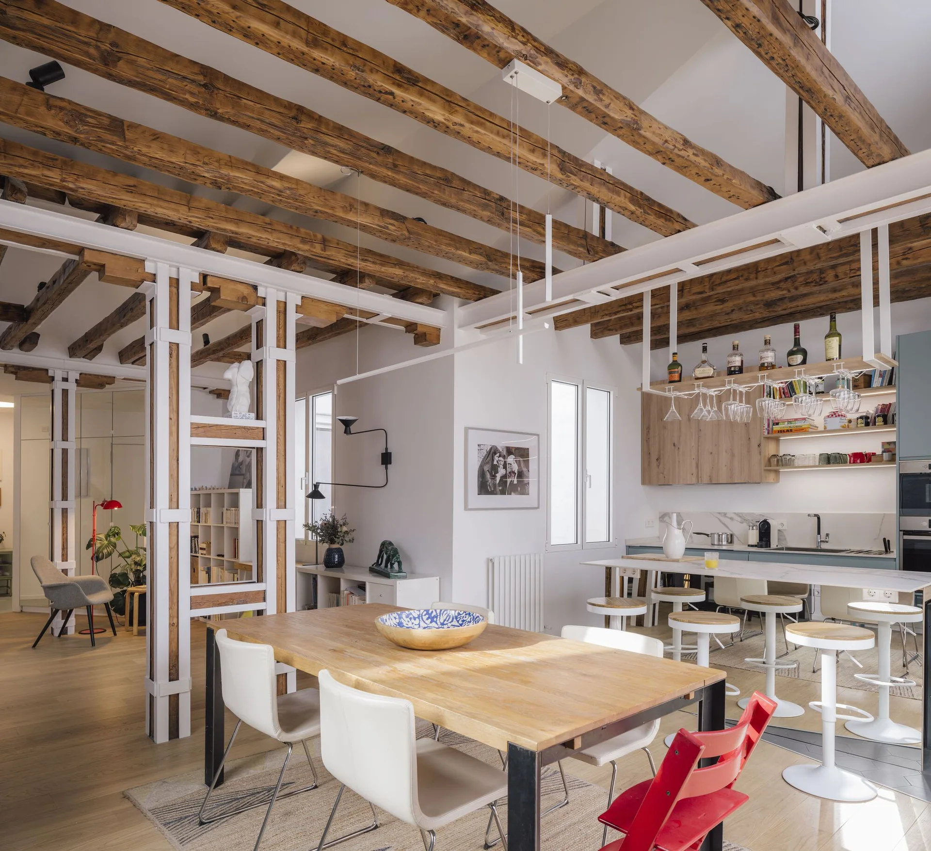 An open plan dining and kitchen area features exposed rustic wood ceiling beams, a wooden dining table, and white architectural room dividers.
