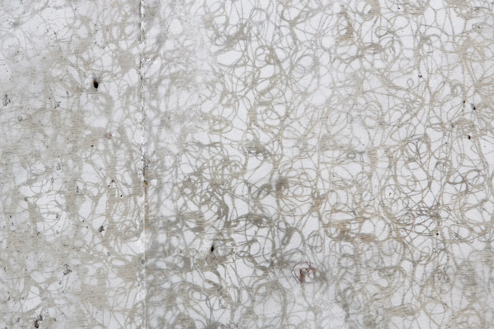 A close-up of a matte grey concrete wall featuring dense, dark swirling scribbles and a subtle vertical joint line on the left.