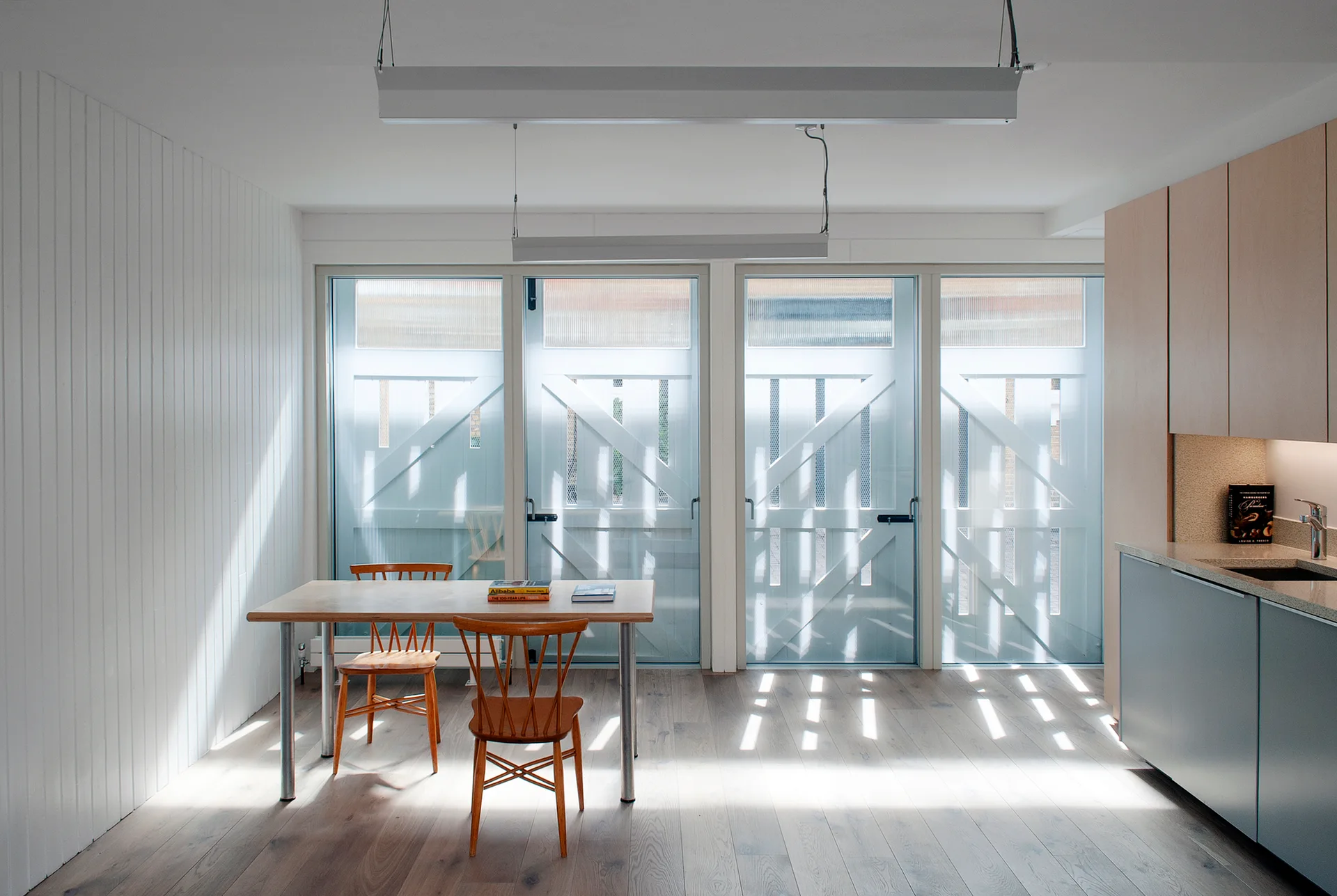 A minimalist kitchen and dining space features a wood table with spindle chairs, a white slatted accent wall, and glass patio doors.