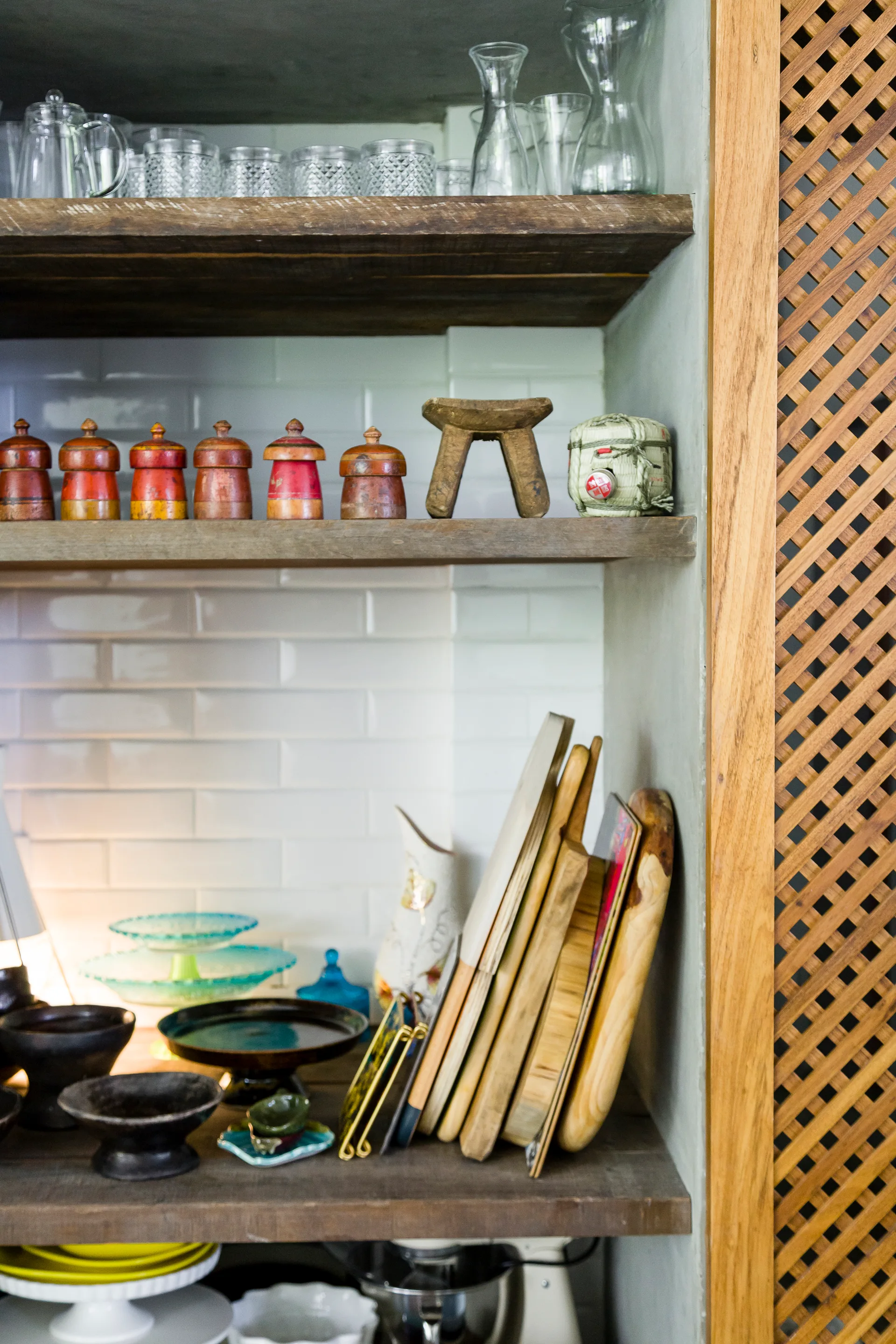 Rustic wooden shelves displaying glassware, red decorative jars, and vertically stacked cutting boards against a white subway tile backsplash.