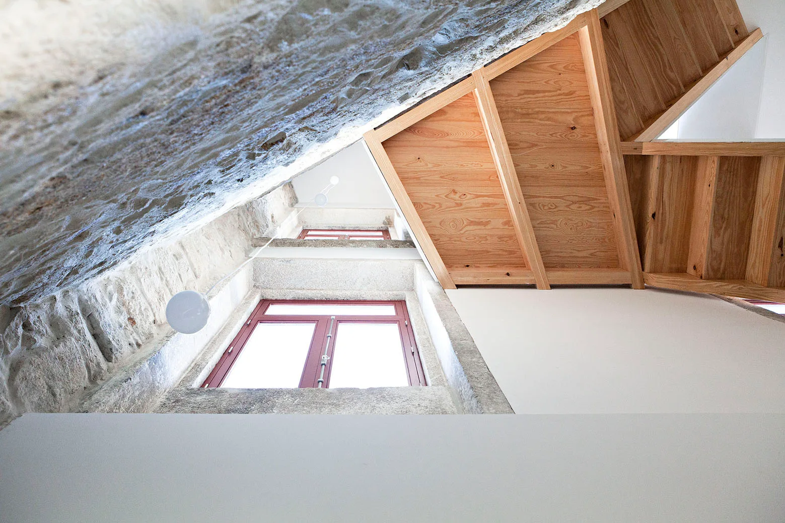 A low-angle view looking up a wooden staircase with exposed stone and white plaster walls next to a window with red frames.