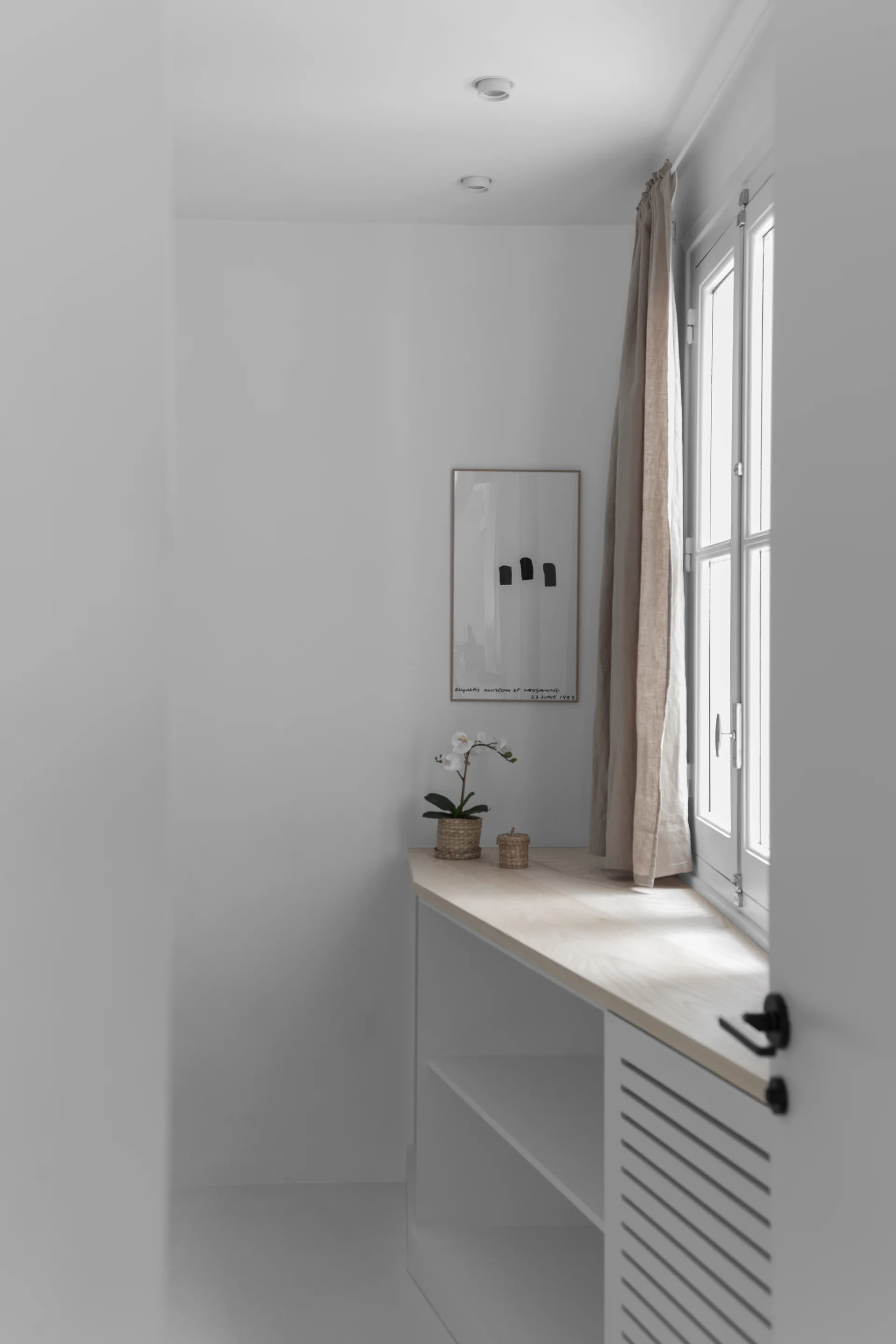 A minimalist white alcove featuring a built-in white cabinet with a light wood top beneath a window dressed with a beige curtain.