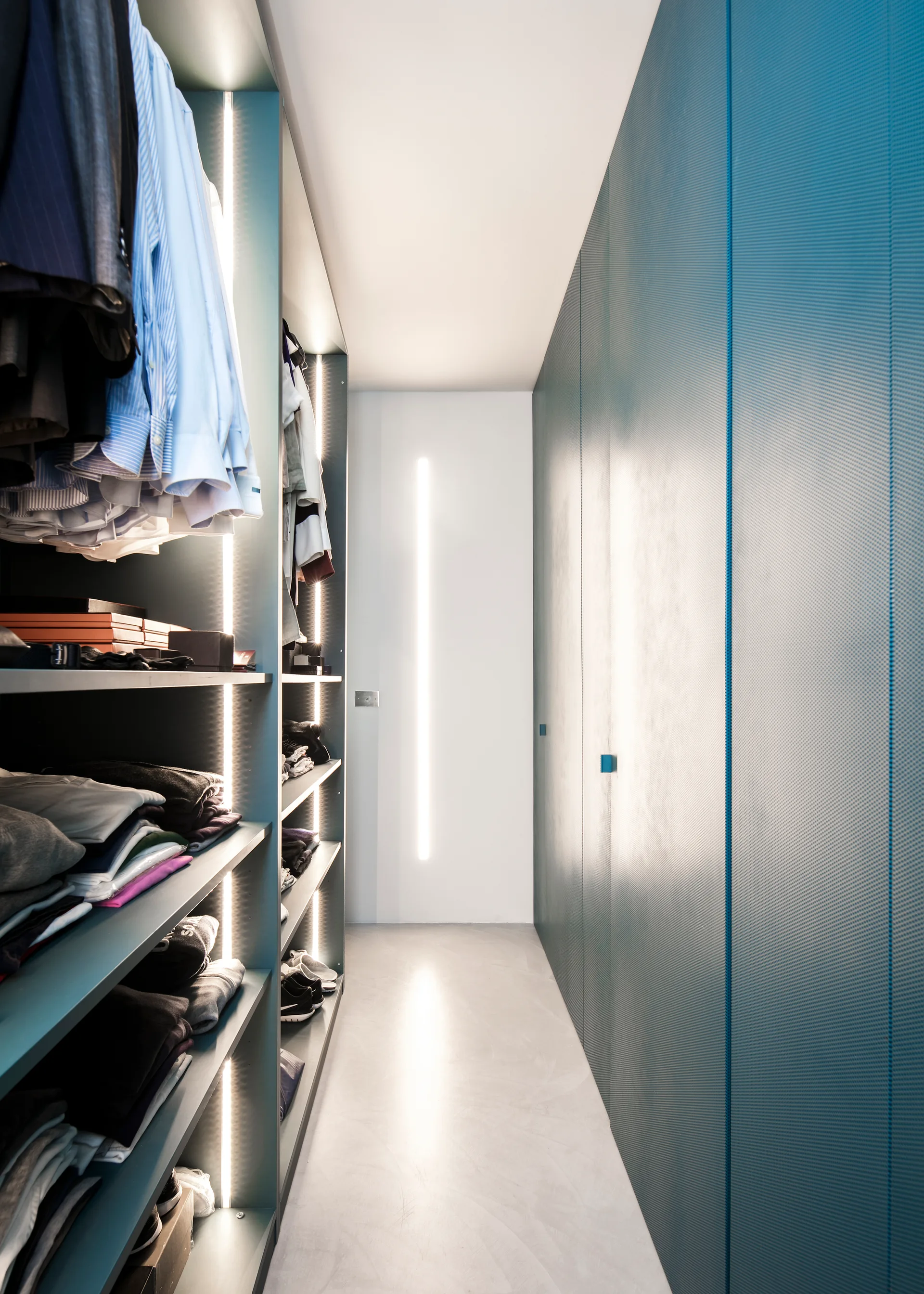 A narrow walk-in closet featuring open teal shelving with vertical LED light strips on the left and tall perforated teal doors on the right.