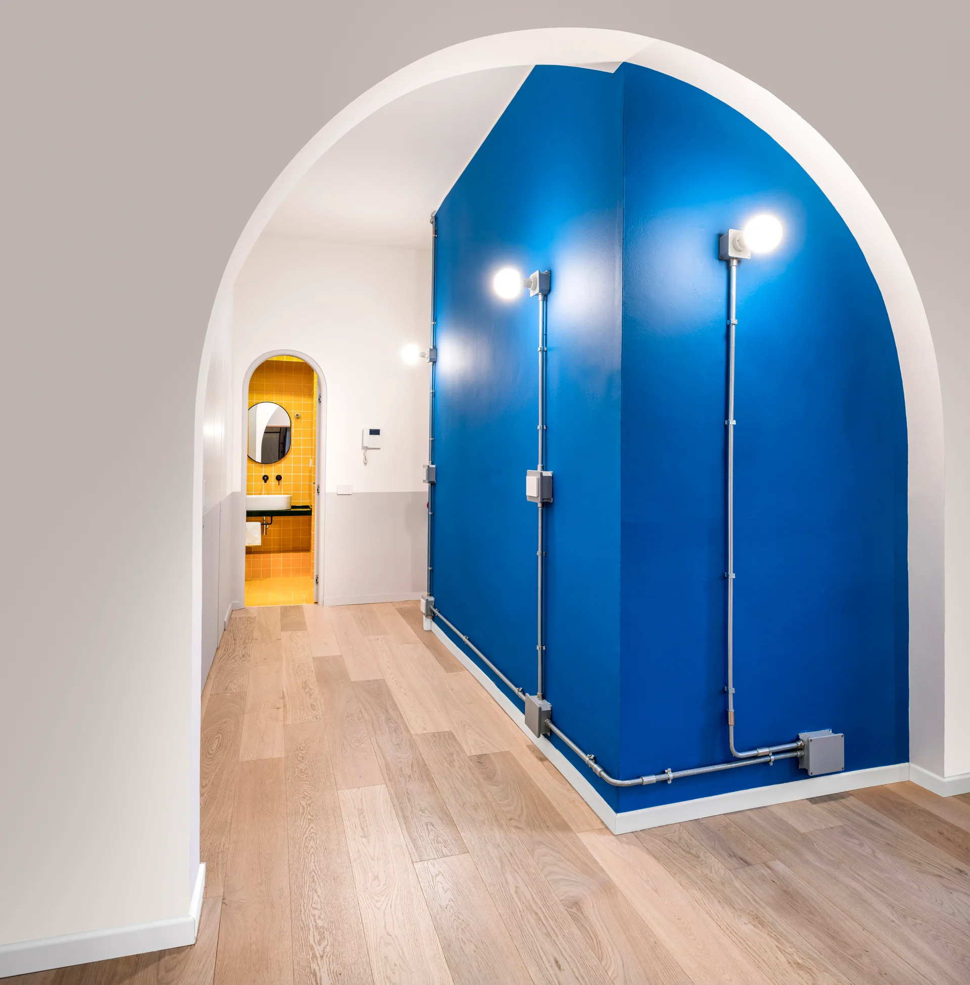 A hallway view through a white archway showing a bright blue wall with exposed metal conduit lighting and a glimpse of a yellow-tiled bathroom.