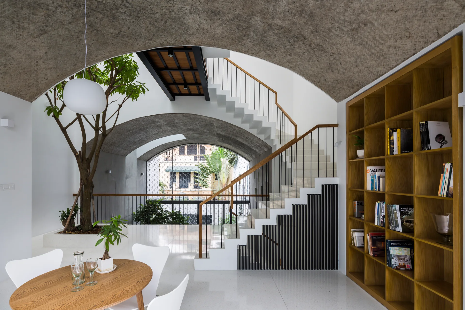 A modern architectural home features a white staircase, a built-in wooden bookcase, concrete vaulted ceilings, and a mature indoor tree.
