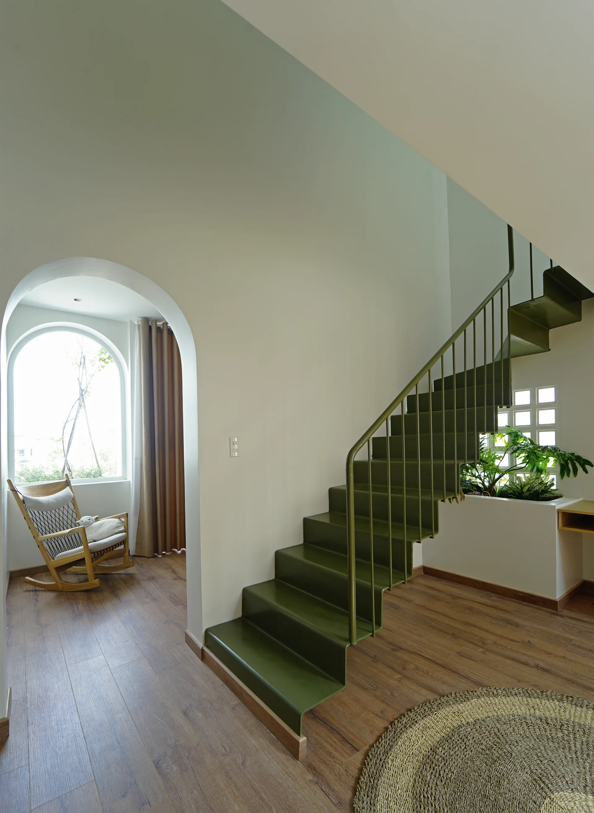 An olive green metal staircase stands next to an arched entryway leading to a sunlit nook with a wooden rocking chair.