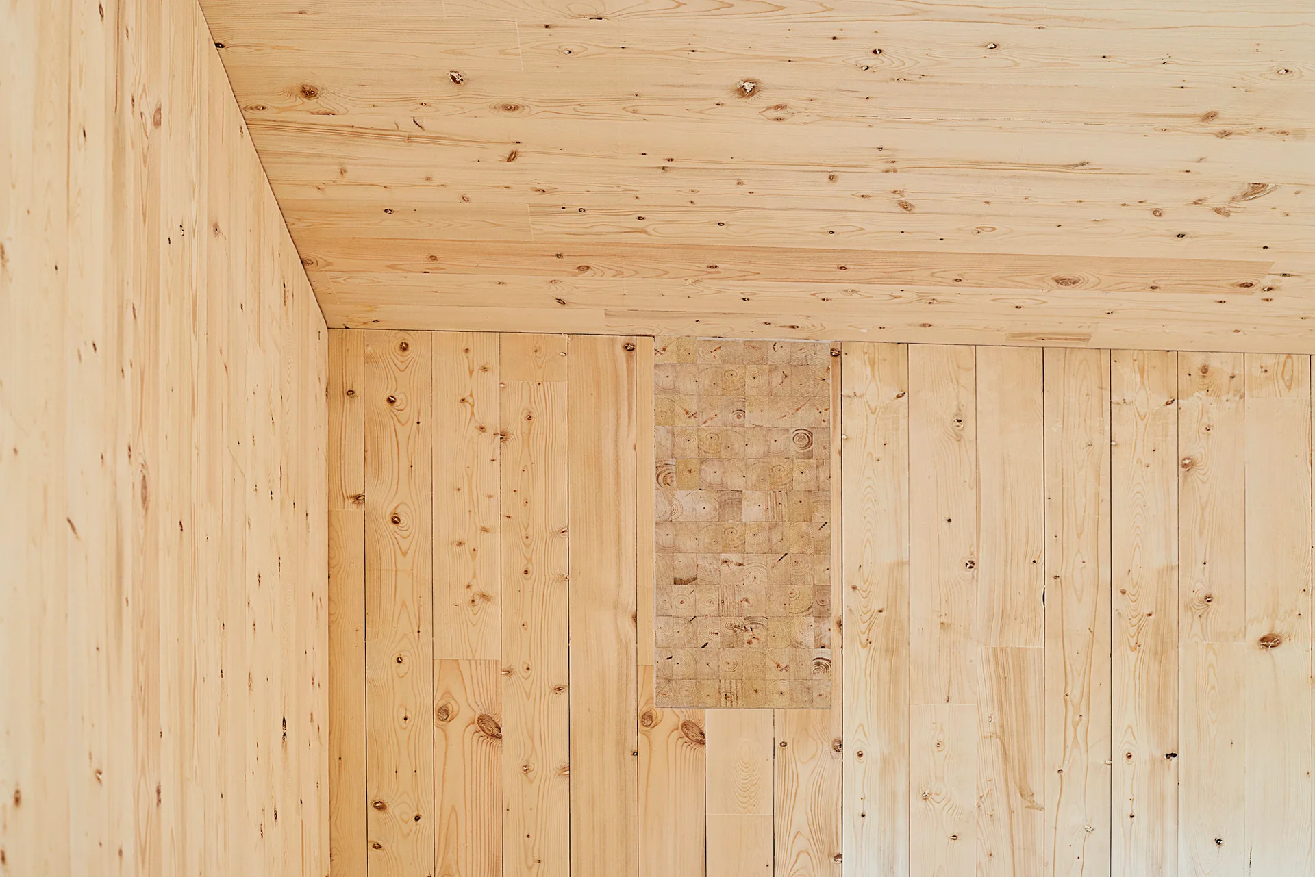 An interior corner with light-toned pine wood plank walls and ceiling featuring a rectangular decorative inlay of square end-grain blocks.