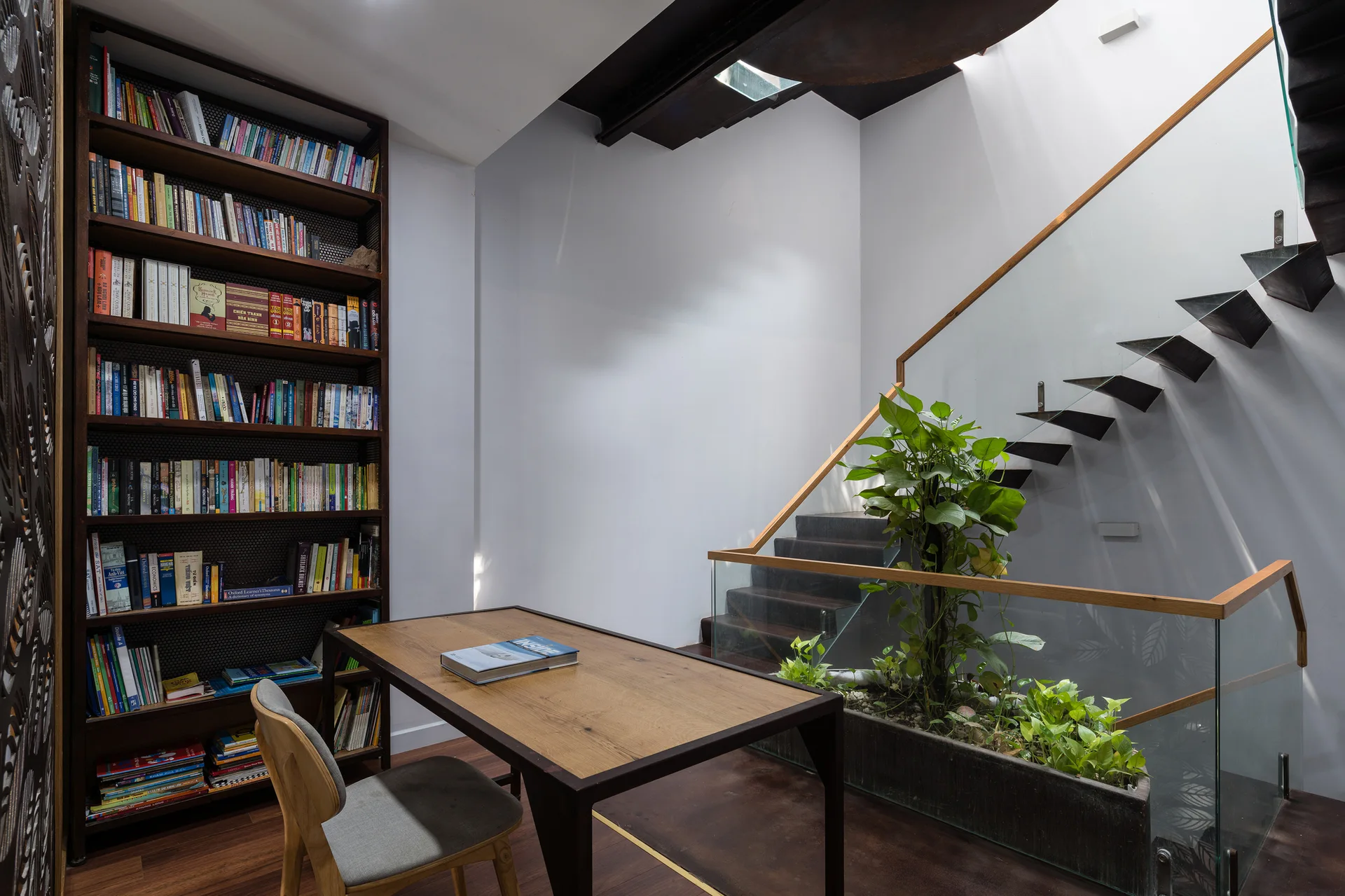 A wooden desk and tall bookshelf sit adjacent to a floating staircase with glass railings and a long built-in indoor planter box.