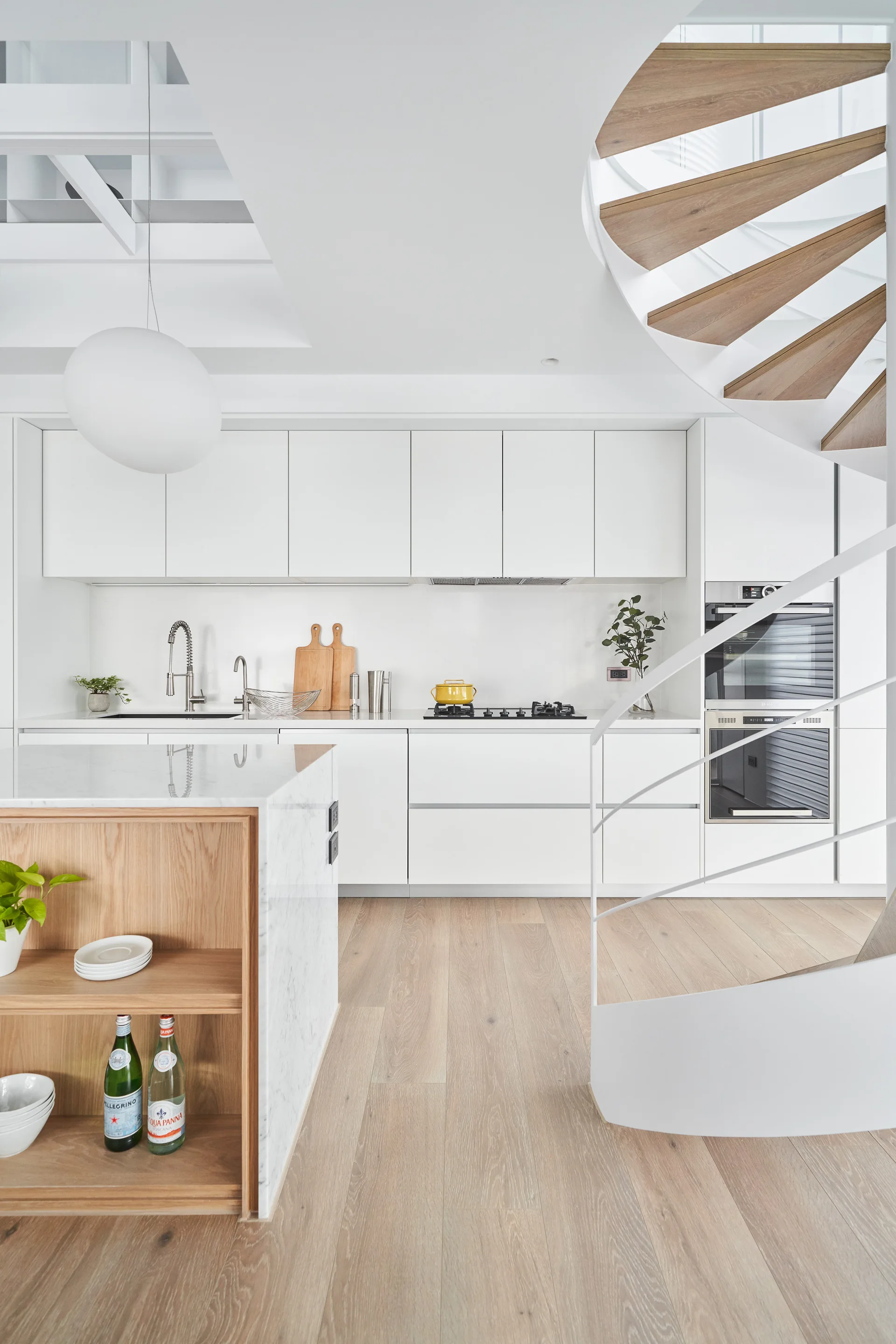 A minimalist white kitchen features a marble island with built-in wood shelves, flat-panel cabinets, and a white spiral staircase.