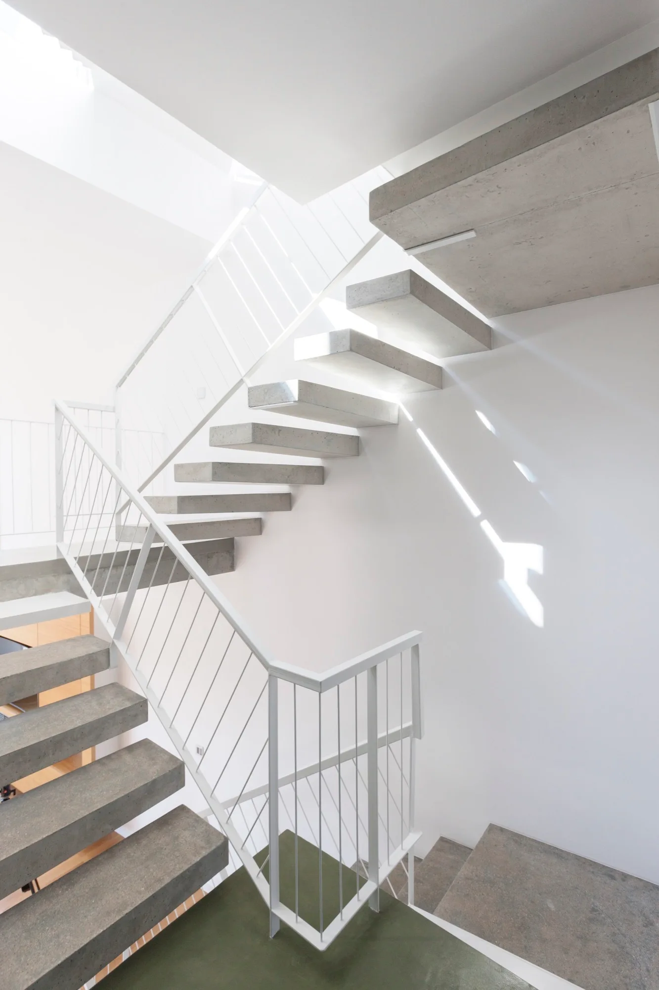 A modern floating staircase with cantilevered concrete treads and a thin white metal railing stands against a plain white wall.