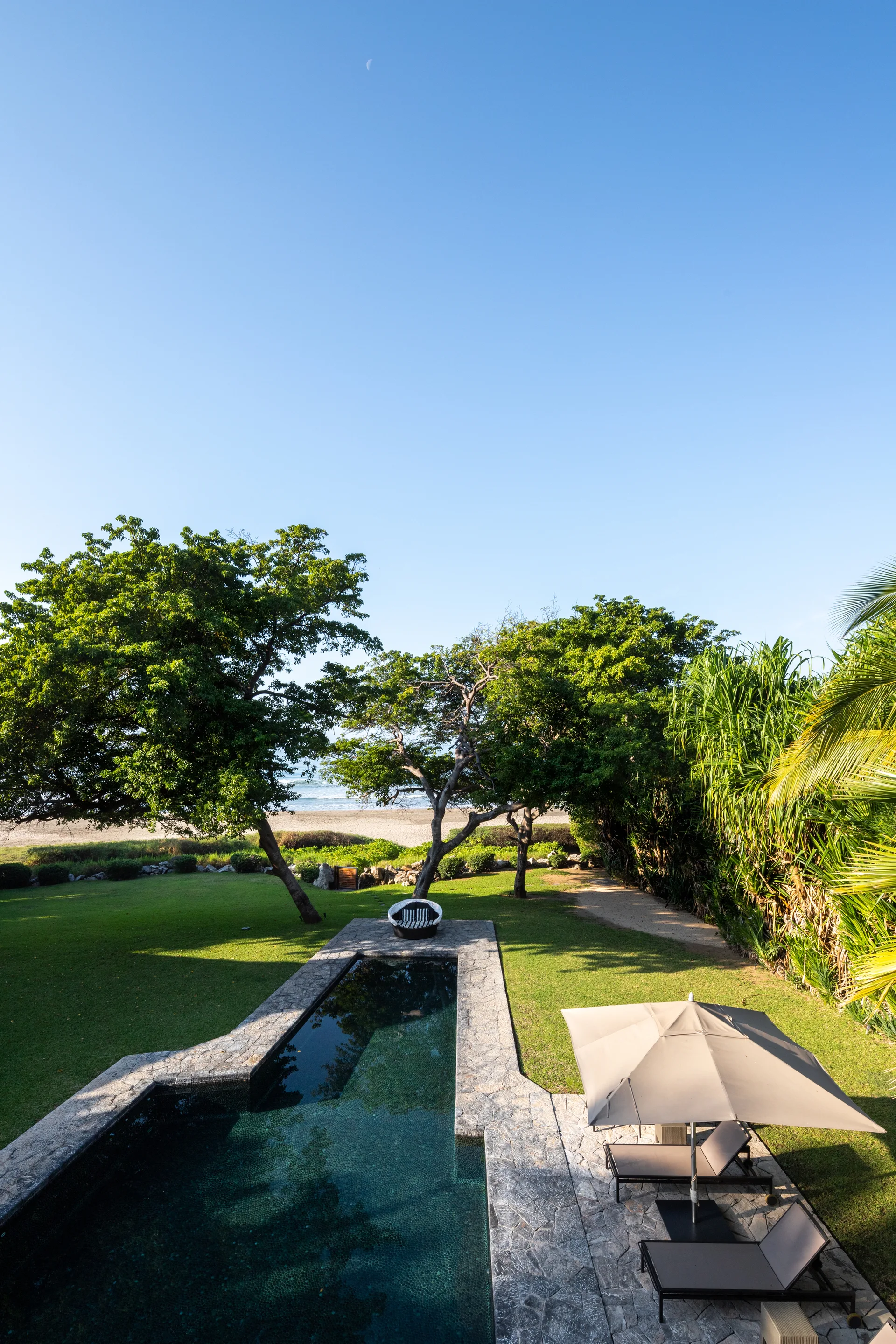 A backyard swimming pool with a grey stone deck is surrounded by a grassy lawn, sun loungers, and mature trees overlooking a distant beach.