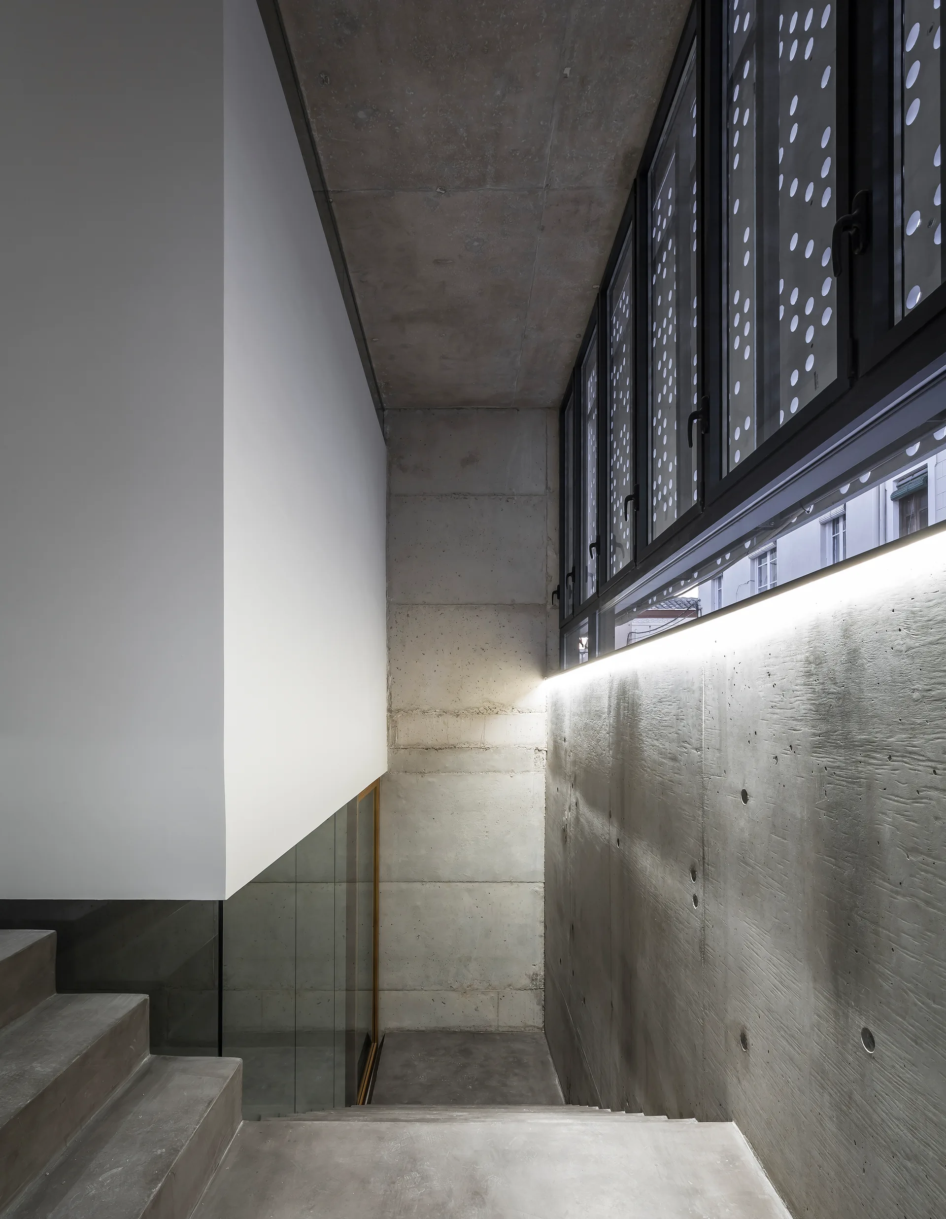 A modern industrial staircase features raw concrete steps and walls illuminated by a long linear LED light strip beneath perforated metal windows.