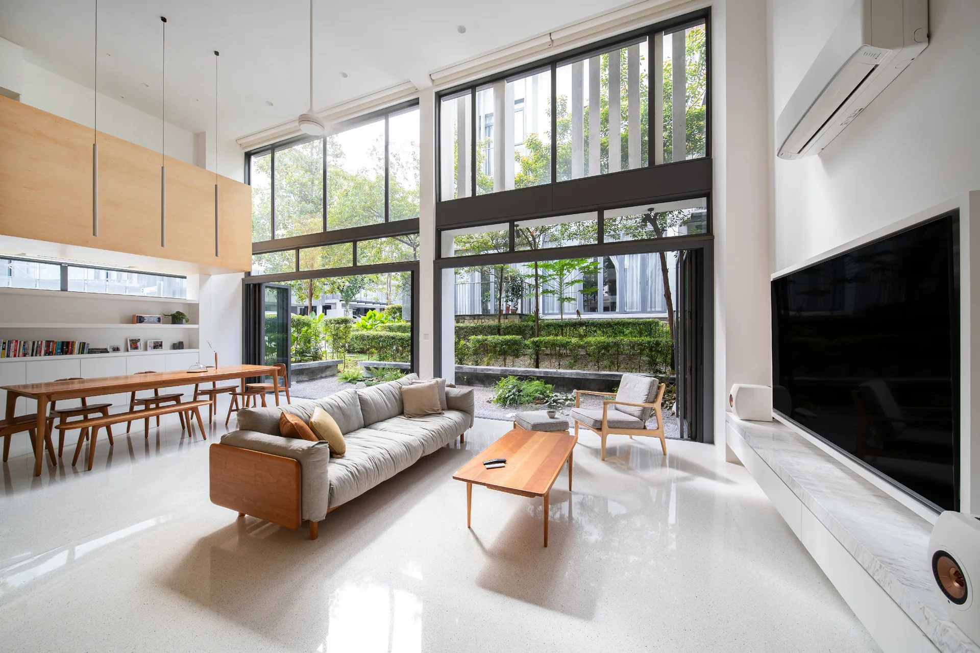 An open-concept living space features a gray sofa, wood dining table, and double-height folding glass doors opening to a lush garden.