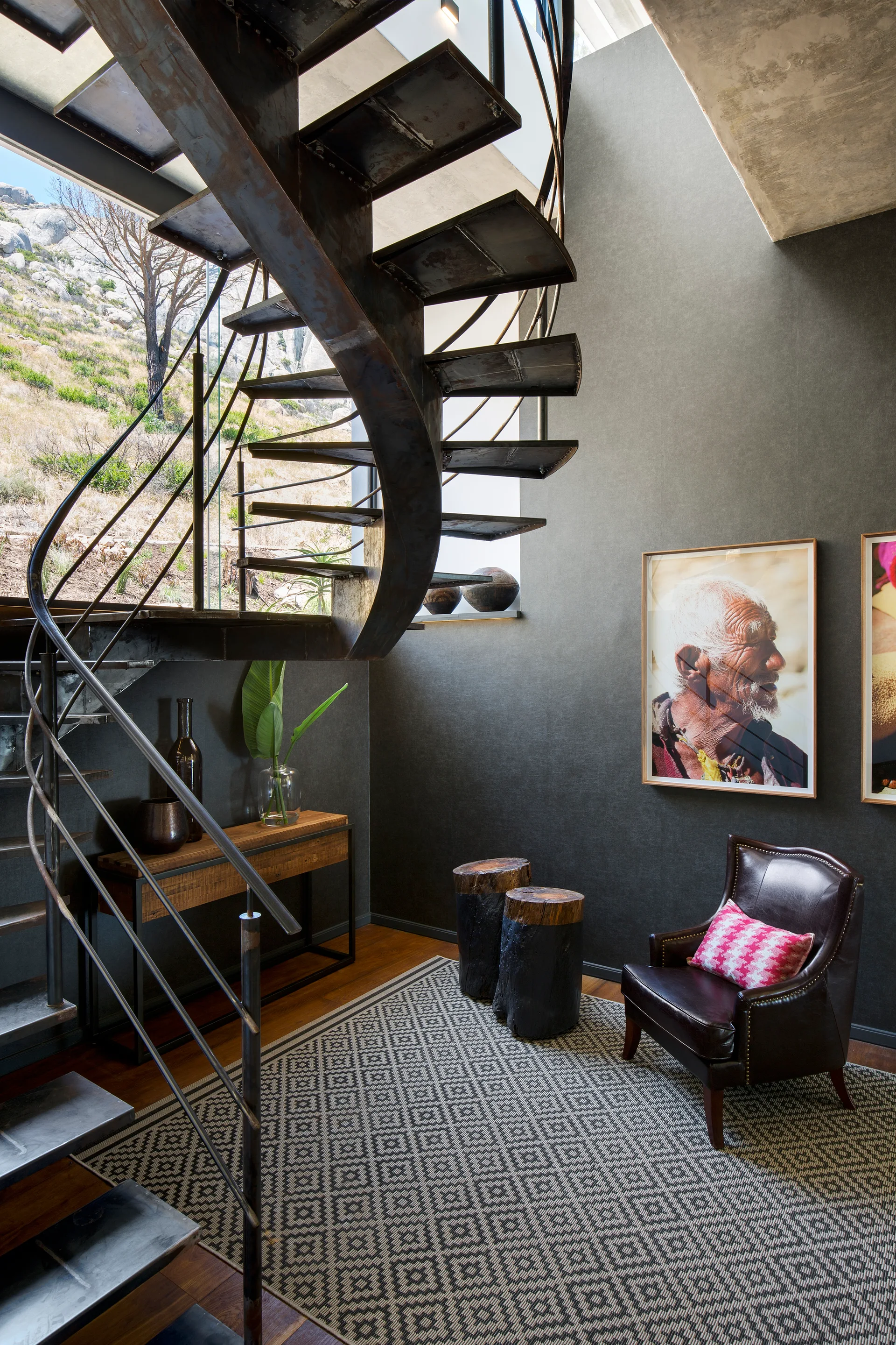 A dark metal spiral staircase curves above a seating area featuring a leather armchair, geometric rug, and stump side tables against a charcoal wall.