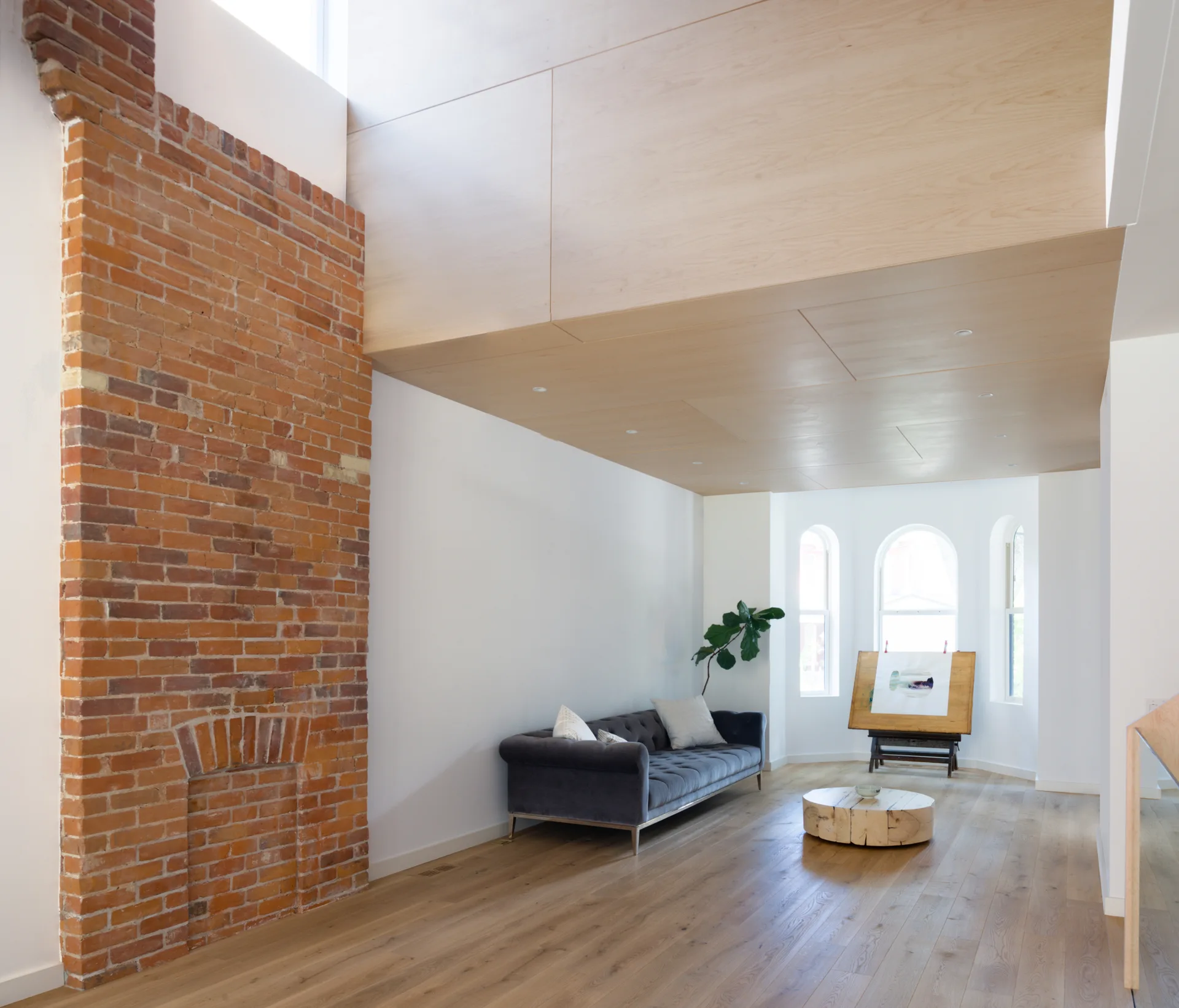 Spacious living room featuring an exposed red brick accent wall, a gray velvet tufted sofa, light wood floors, and a wood paneled ceiling.
