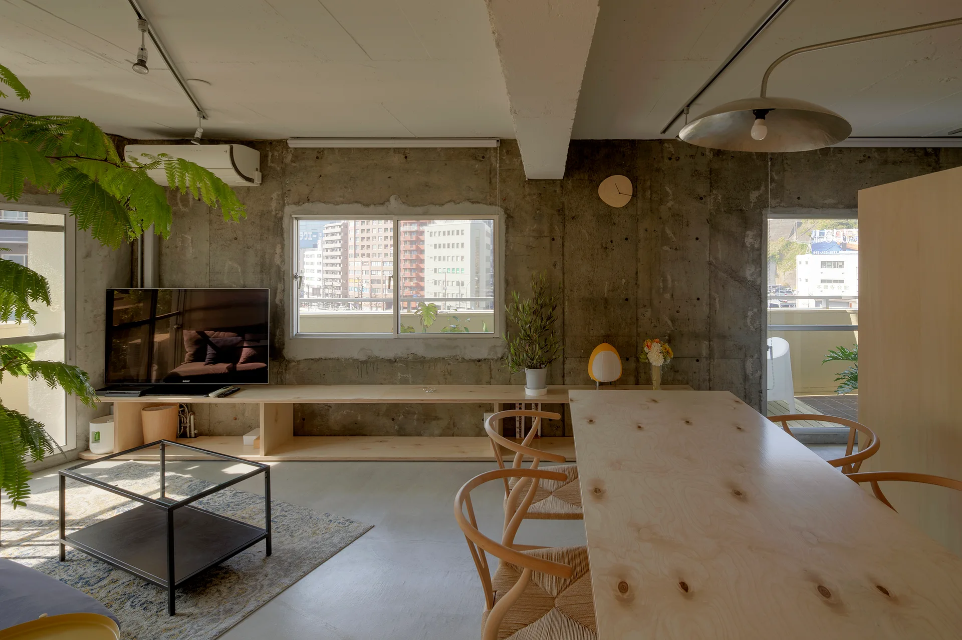An open-concept living and dining area featuring raw concrete walls, a plywood dining table with matching chairs, and a low media console.