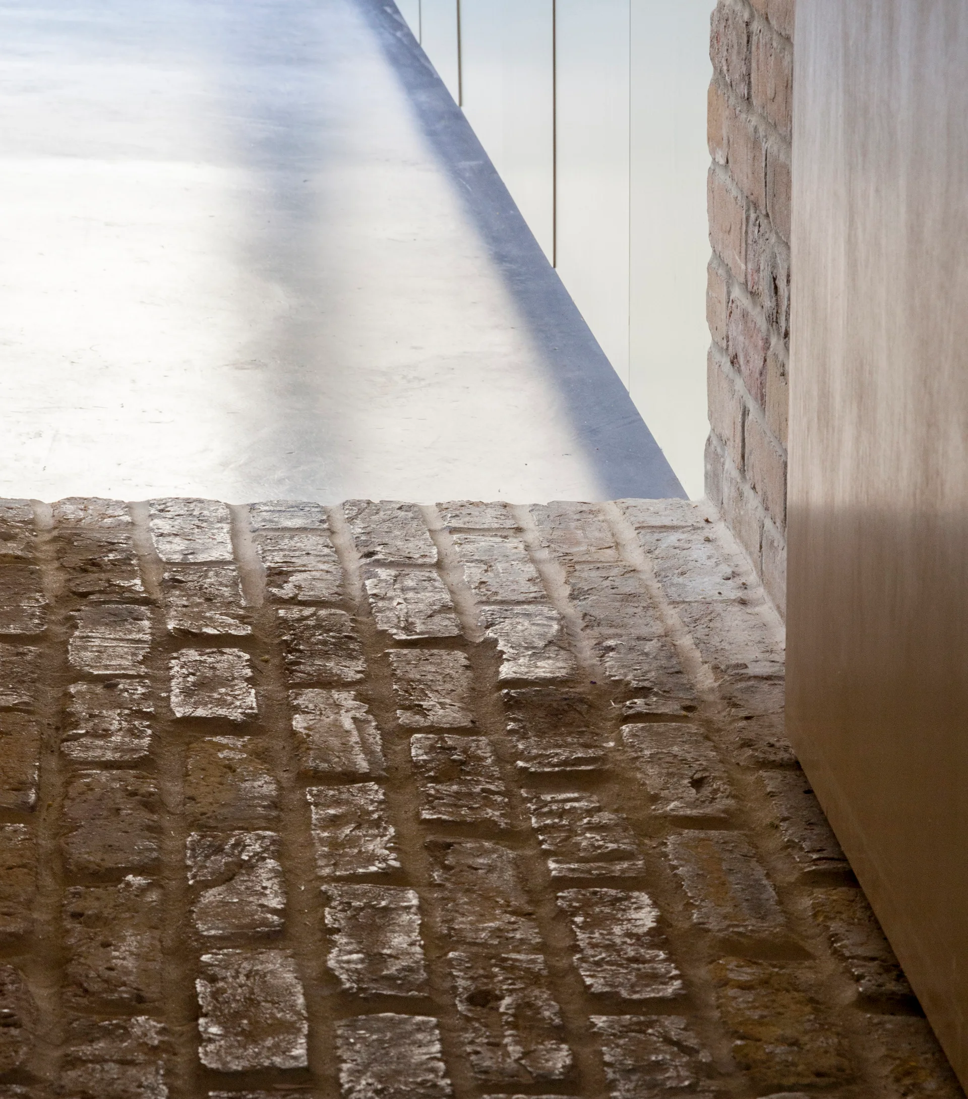 A close-up of an interior floor transition where rough, textured brown brick meets a smooth light grey floor next to a white wall panel.