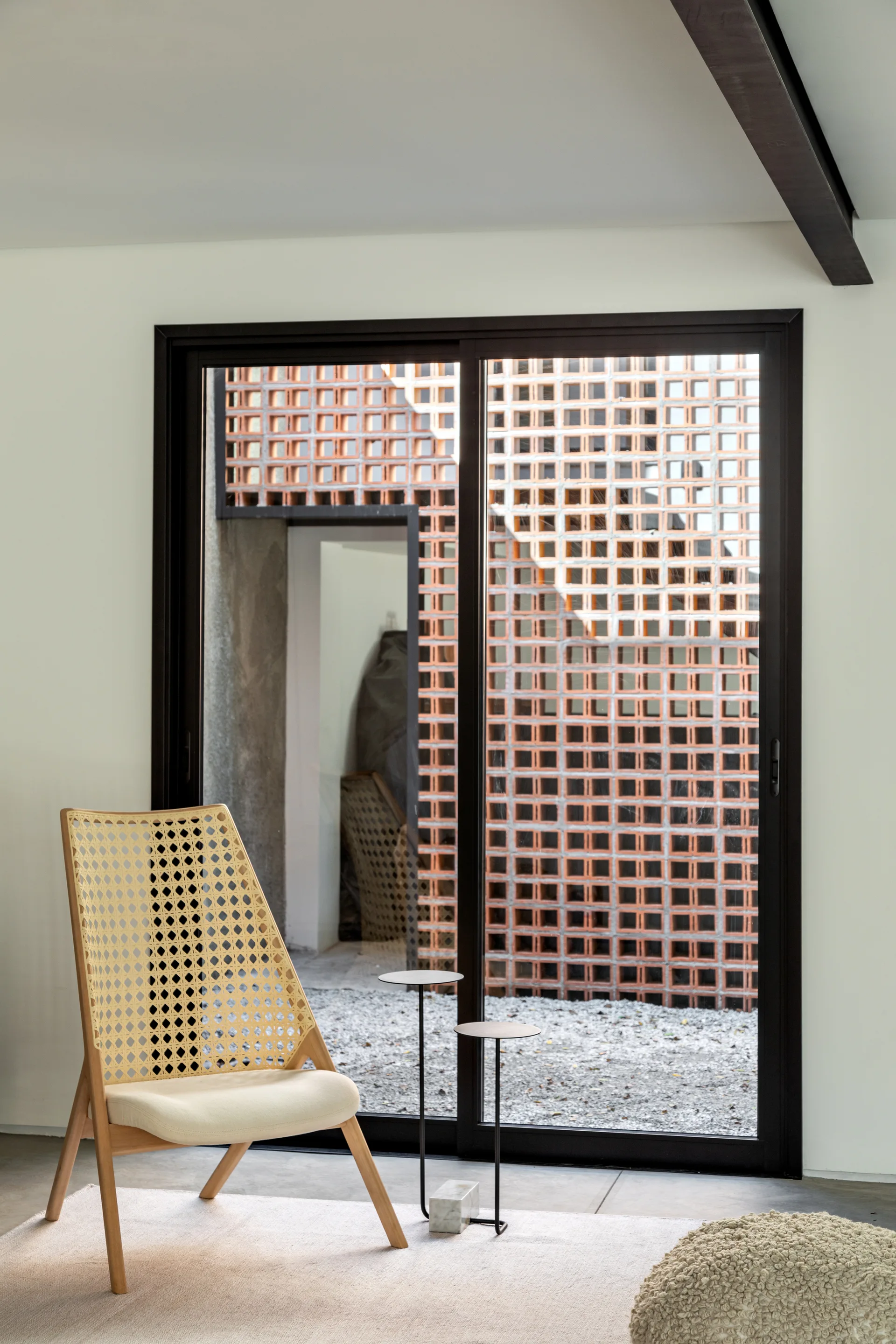 A woven rattan accent chair and thin metal side tables sit on a light rug in front of a sliding glass door looking out at a terracotta breeze block wall.