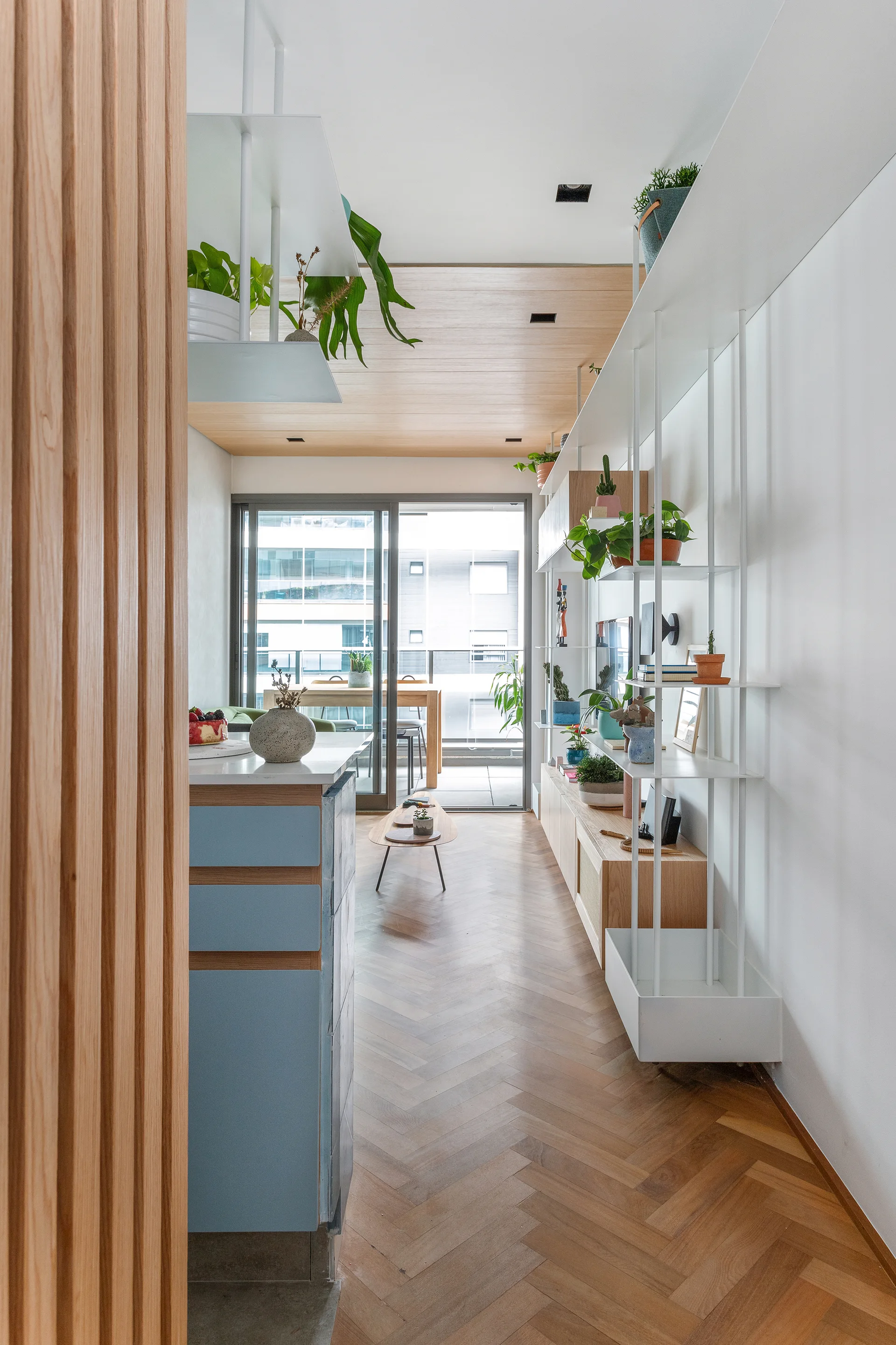 A modern apartment interior featuring herringbone wood flooring, a light blue kitchen island, a tall white metal shelving unit, and a balcony view.