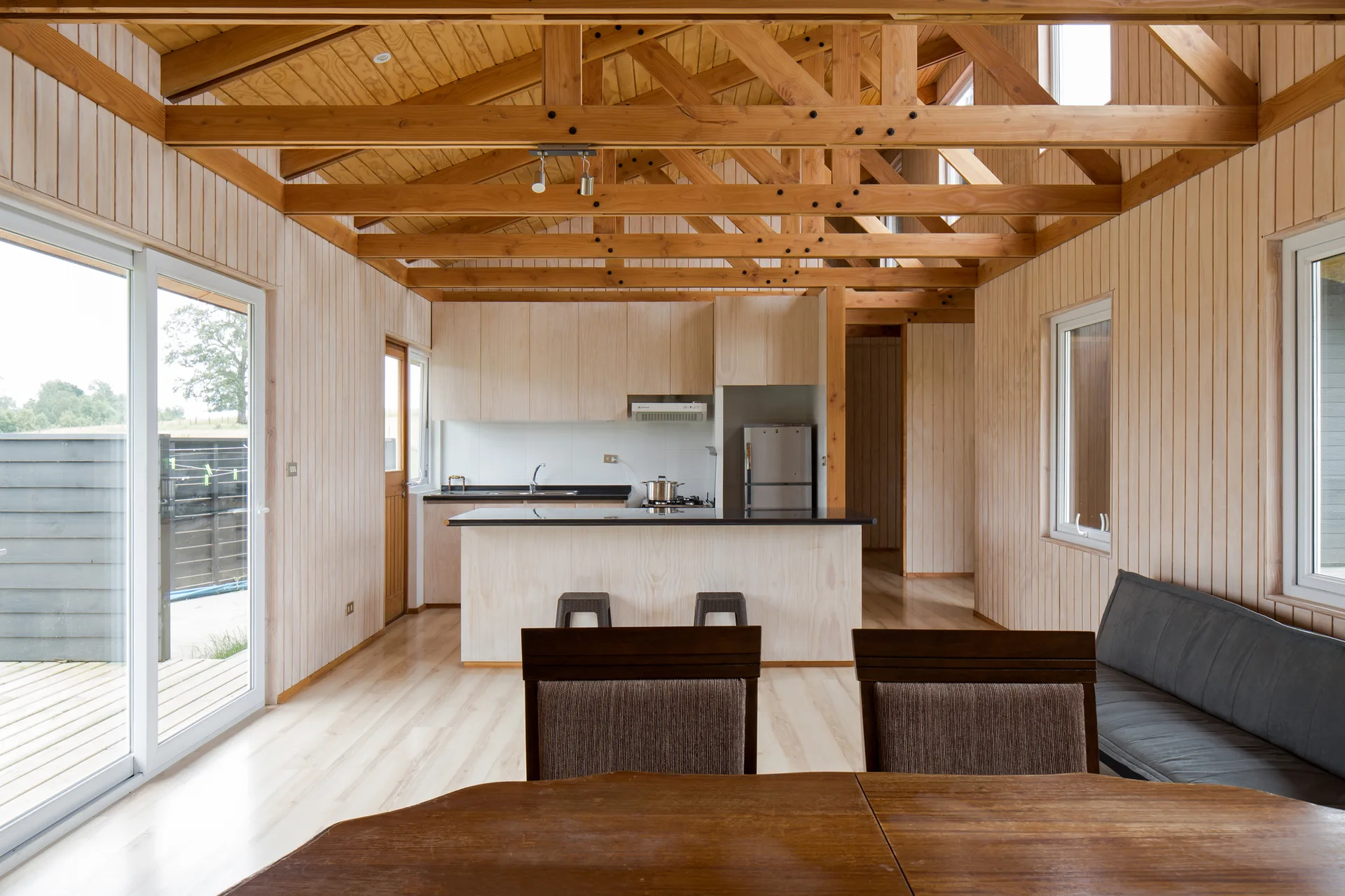 An open-plan cabin kitchen and dining area featuring light wood paneled walls, a vaulted ceiling with exposed timber trusses, and a central island.