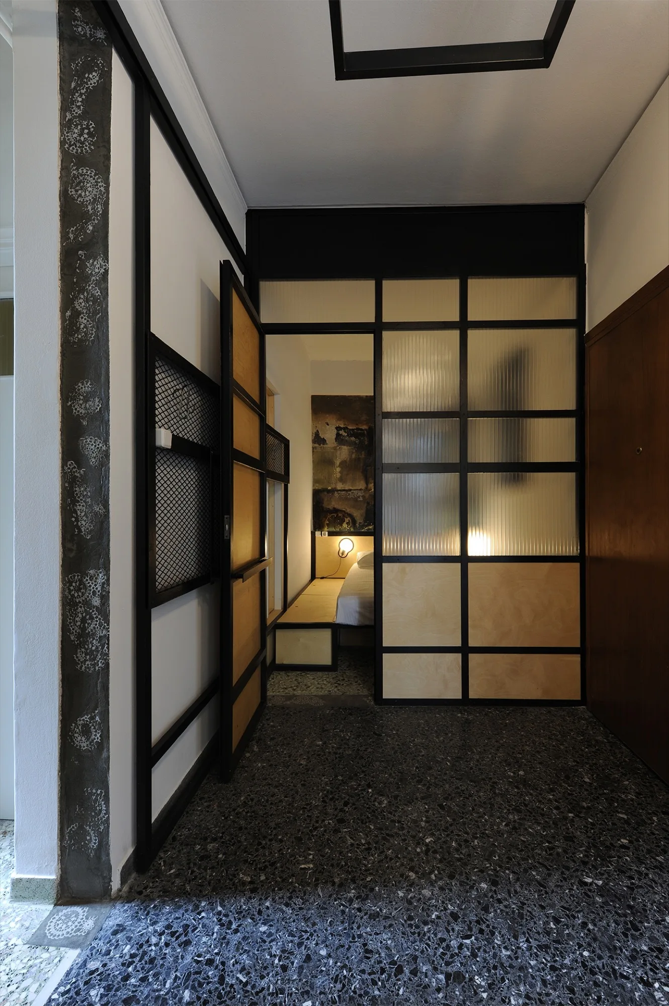 A black metal grid partition with fluted glass and wood panels separates a dark terrazzo entryway from a minimalist bedroom with a platform bed.