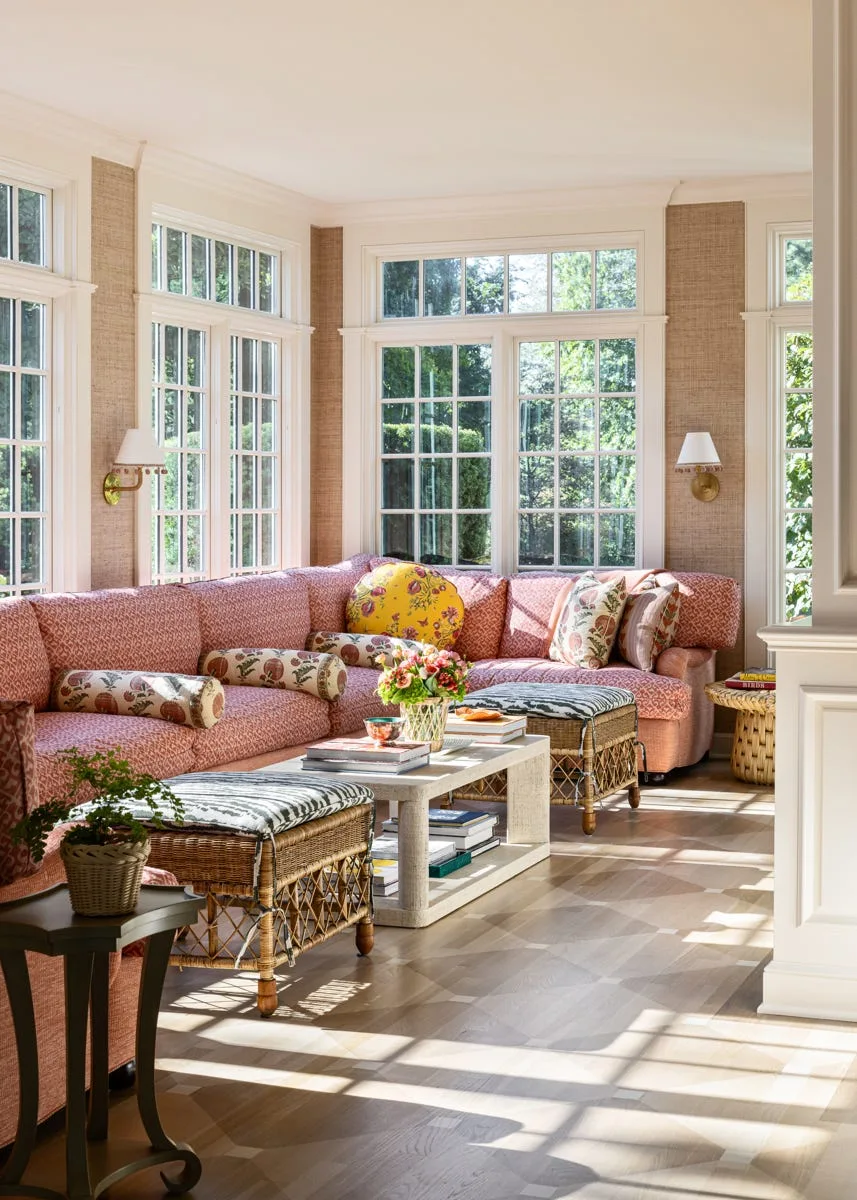 A sunlit sunroom features a large pink patterned sectional sofa, wicker ottomans with animal-print cushions, and tall paneled windows against textured walls.