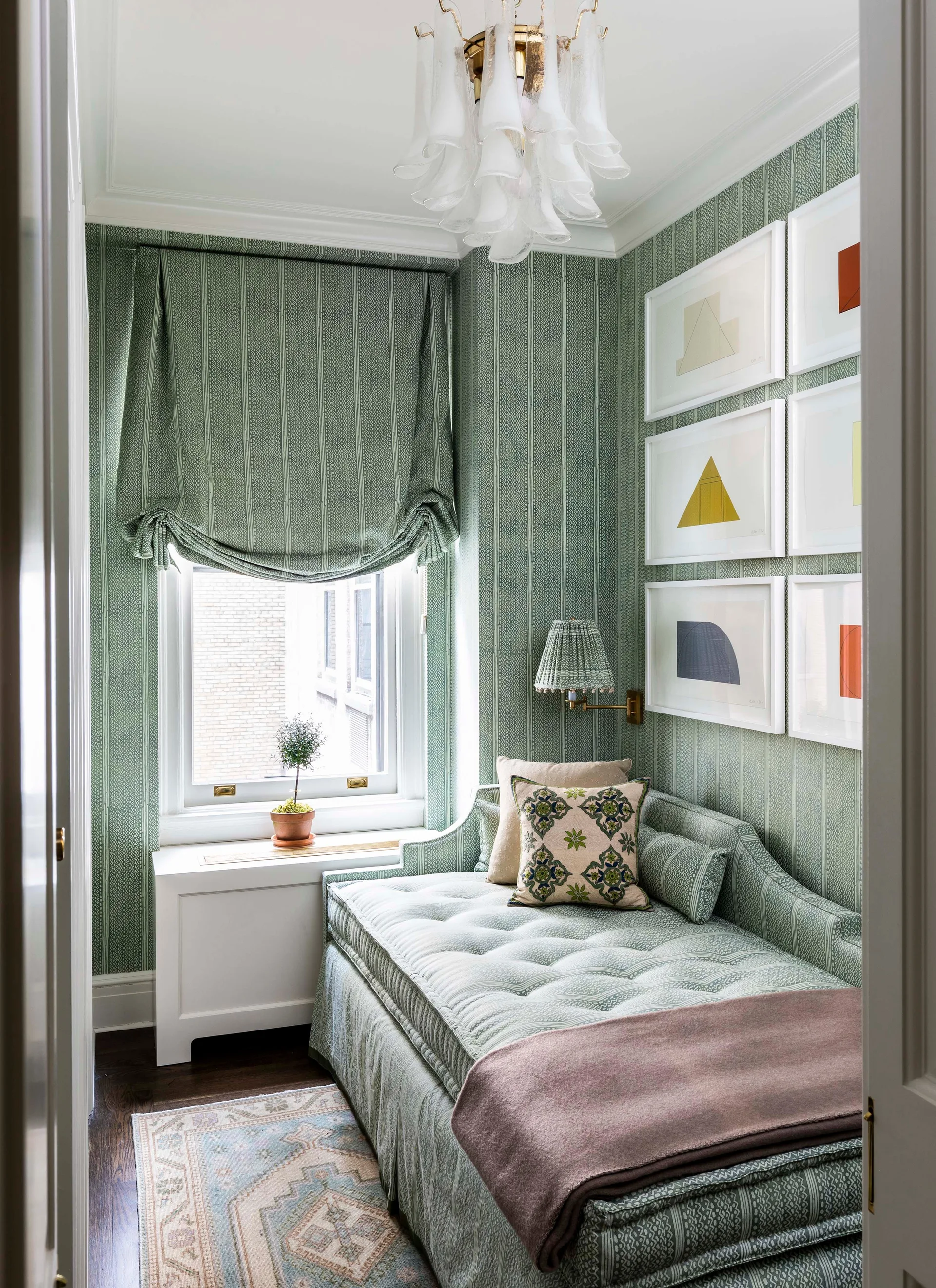 A small bedroom features a tufted daybed and walls wrapped in matching sage green patterned wallpaper beneath a glass chandelier and a gallery wall.