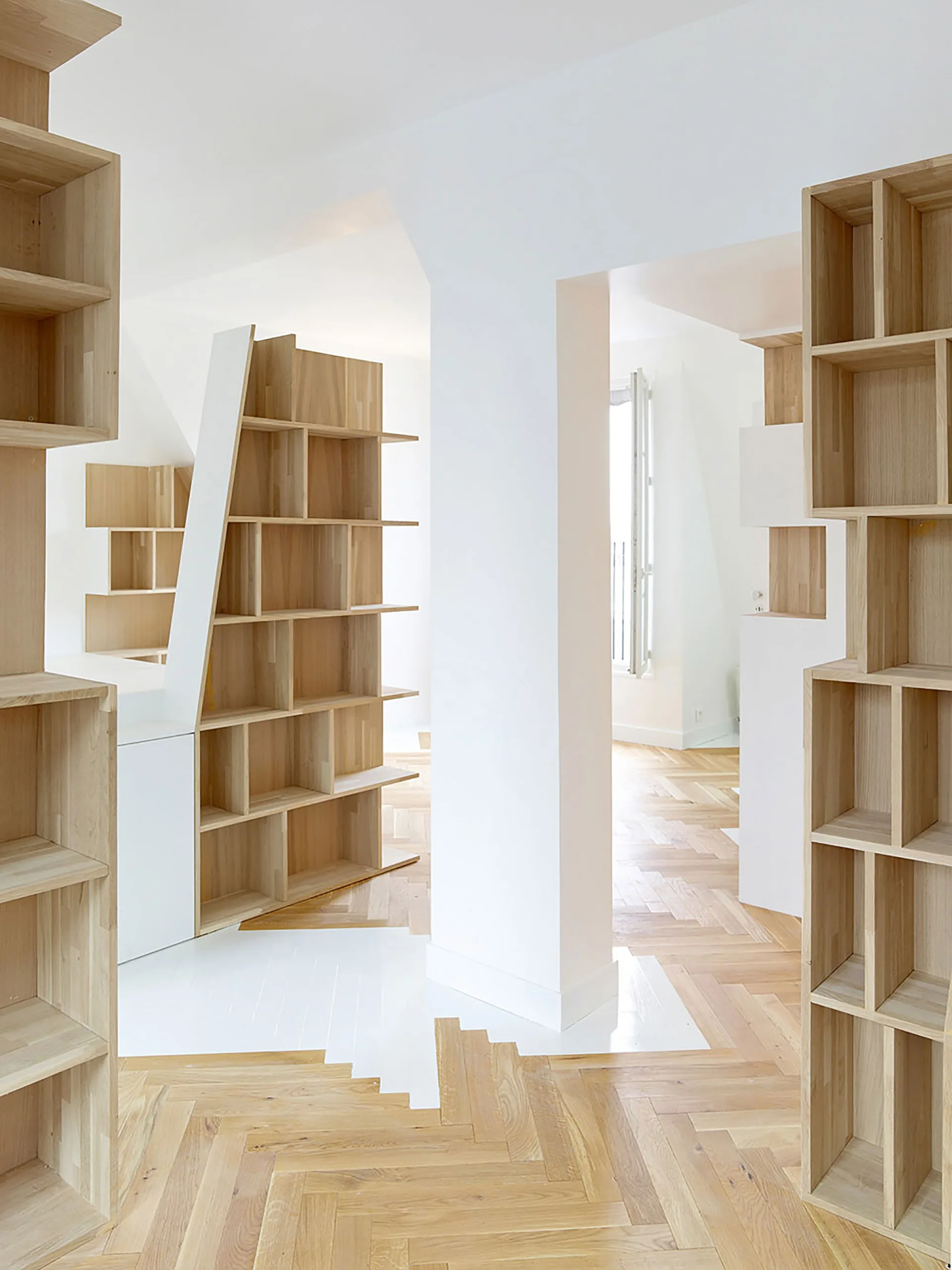 Custom light wood bookshelves stand on a light-colored herringbone wood floor beside a white structural column in a bright interior.