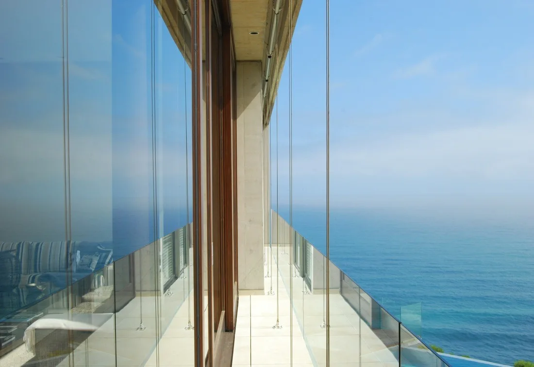 A long outdoor balcony features a frameless glass railing overlooking a blue ocean, adjacent to a sliding glass door with a wood frame.