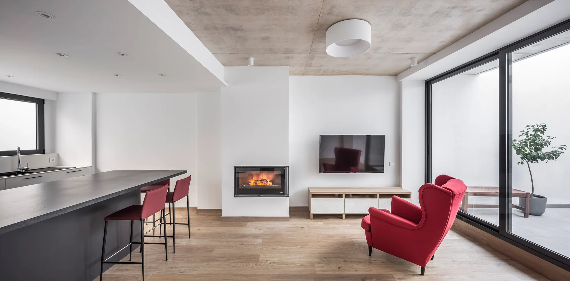 A modern open-plan living room and kitchen features an exposed concrete ceiling, a built-in fireplace, and a bright red armchair.
