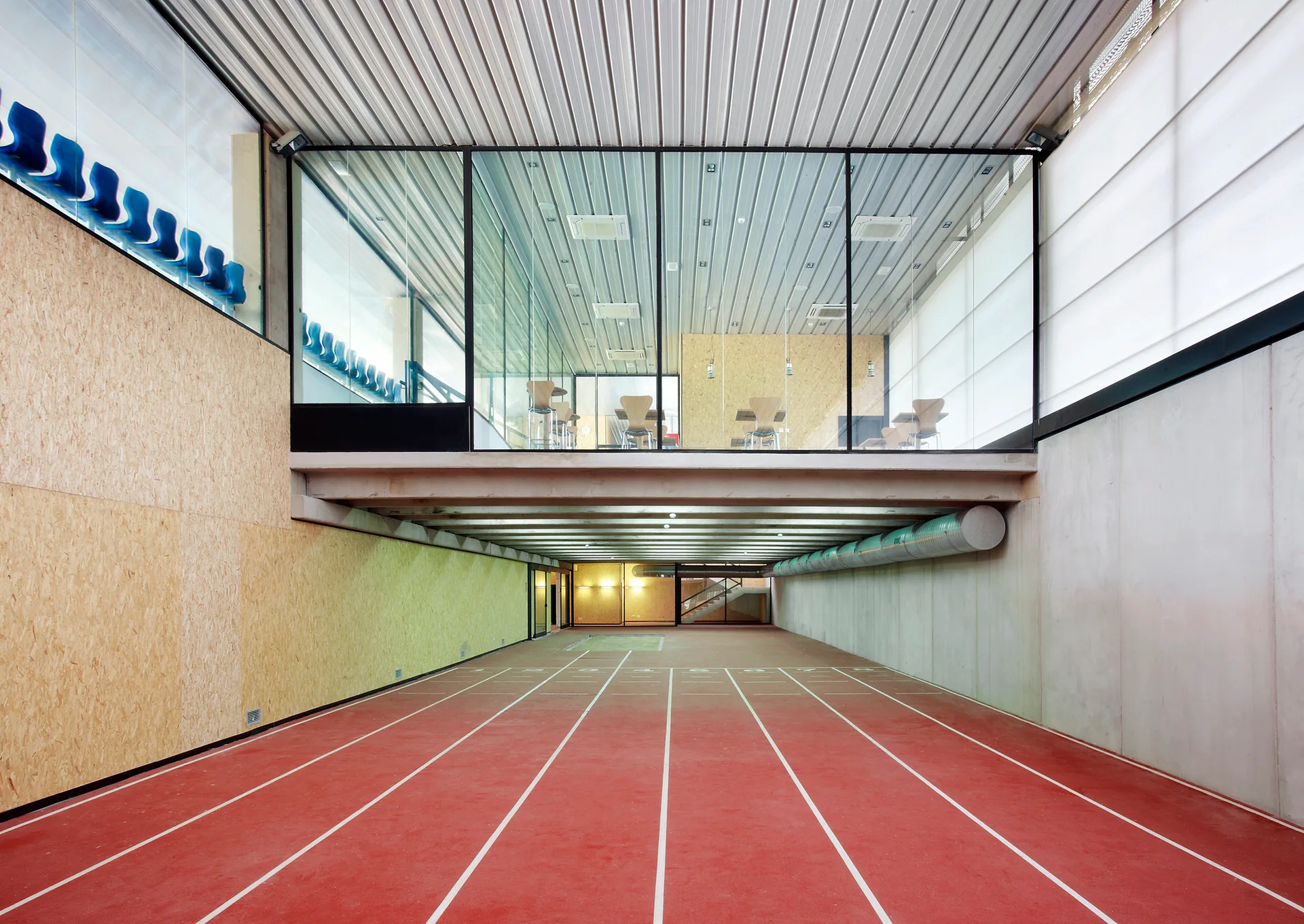 An indoor athletic facility featuring a red running track beneath a glass-walled mezzanine level and textured wood walls.