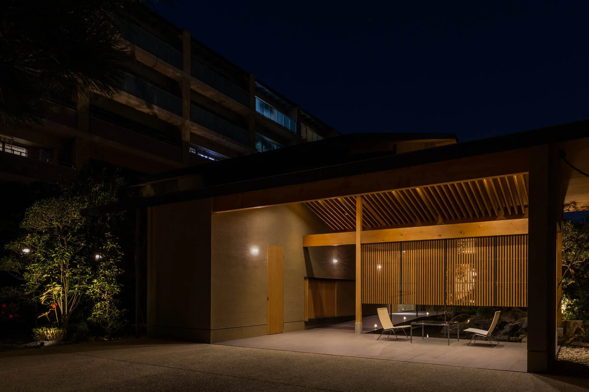 A modern covered outdoor pavilion at night with a slatted wooden screen, exposed ceiling beams, and minimalist patio furniture.