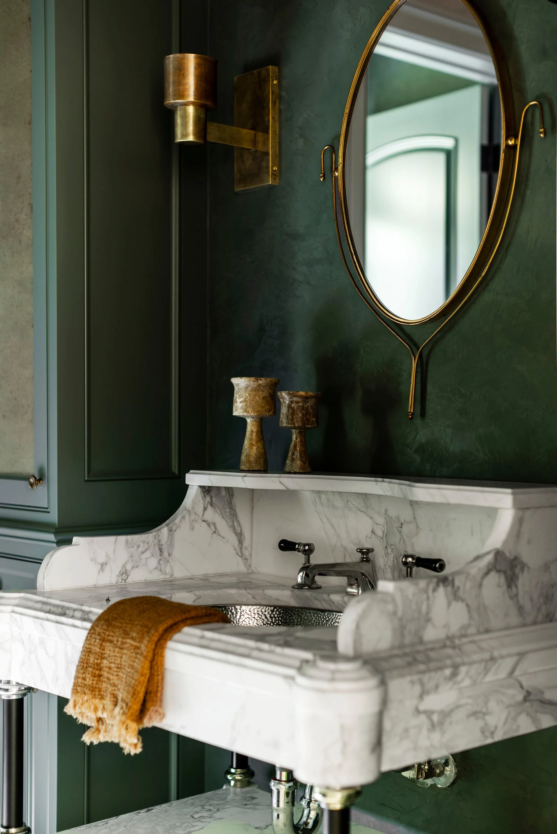 A marble console sink with classic metal fixtures stands against a dark green paneled wall under an oval brass-framed mirror and a brass wall sconce.